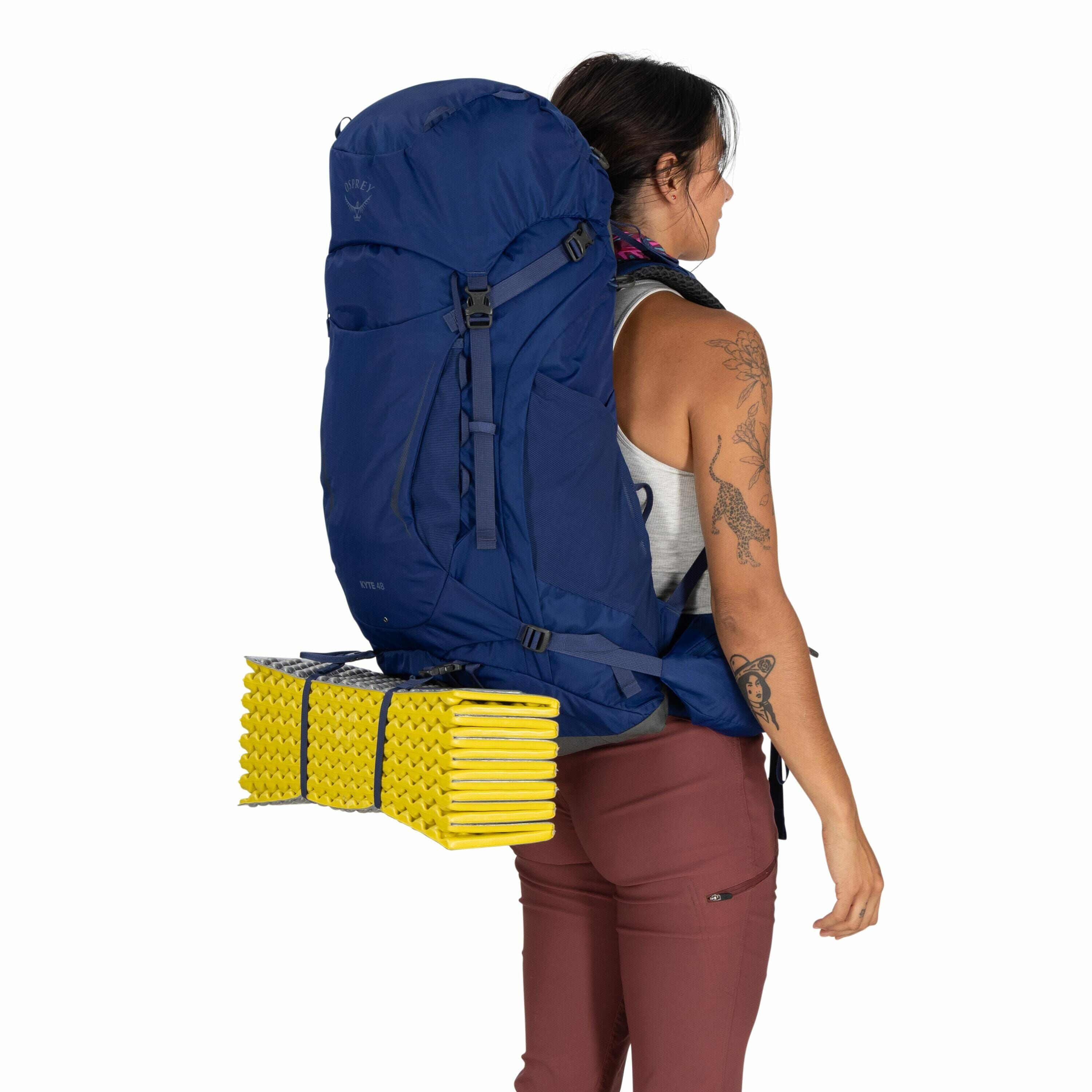 The image features a person wearing a large blue Osprey backpack, which appears well-suited for hiking or backpacking trips. Attached to the bottom of the backpack is a bright yellow, folded sleeping pad, a common accessory for outdoor camping to provide