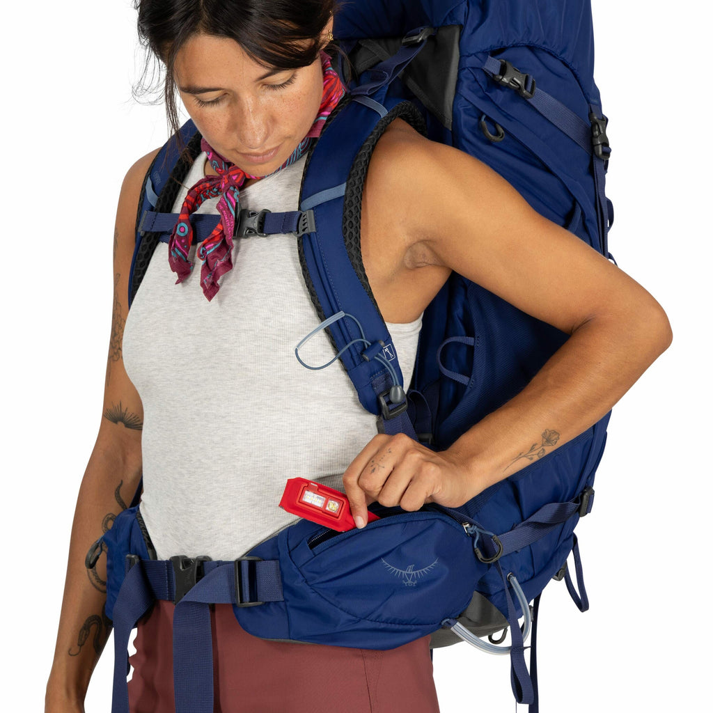 The image shows a person dressed in outdoor attire, carrying a large blue hiking backpack. They are wearing a gray sleeveless top and have a colorful scarf tied around their neck. The backpack features multiple compartments and straps, indicating it's des