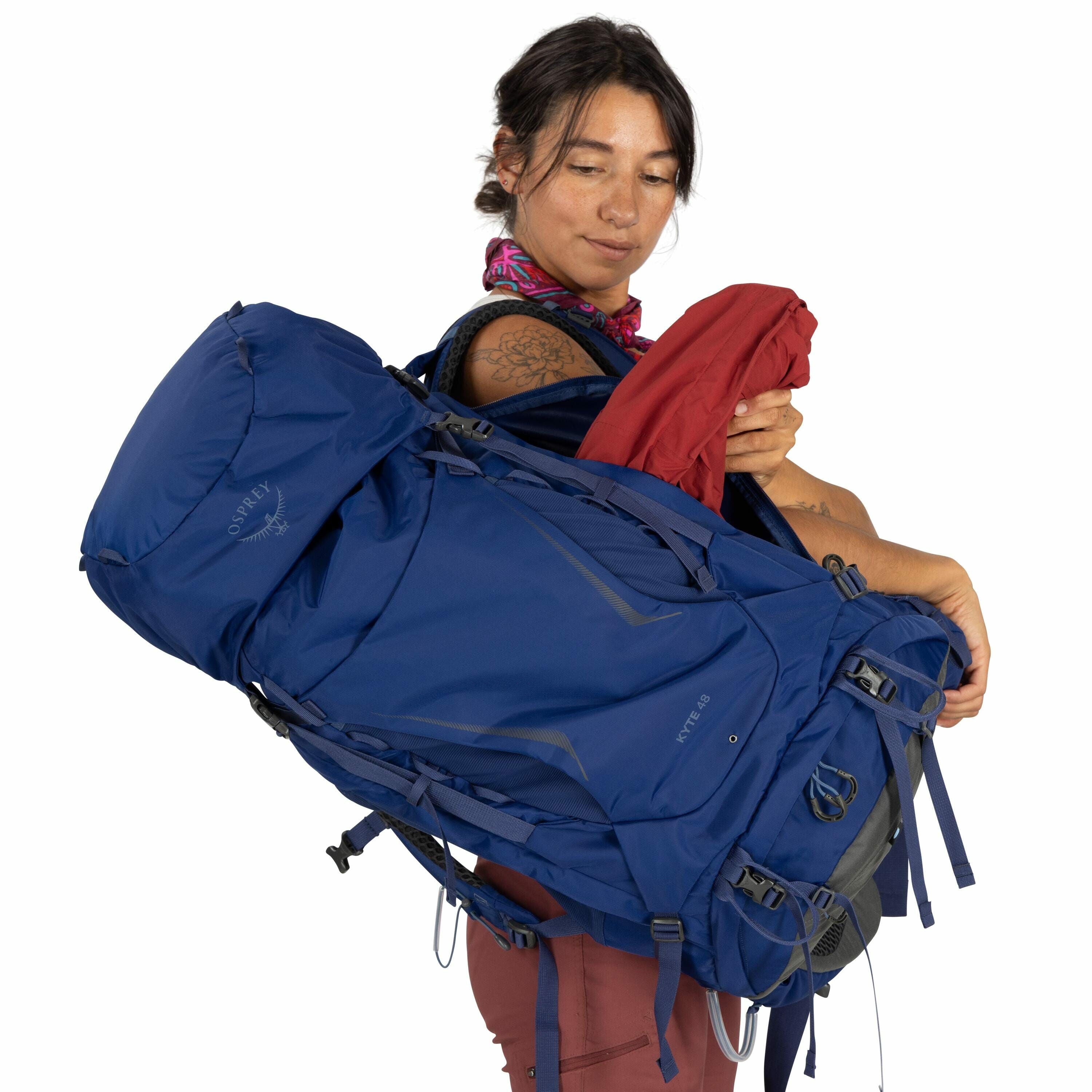 The image shows a person holding a large blue Osprey backpack with the model name "Kyte 46" visible on the side. The backpack features numerous straps and buckles designed for adjustable fit and secure storage, indicating it's meant for hiking or travelin