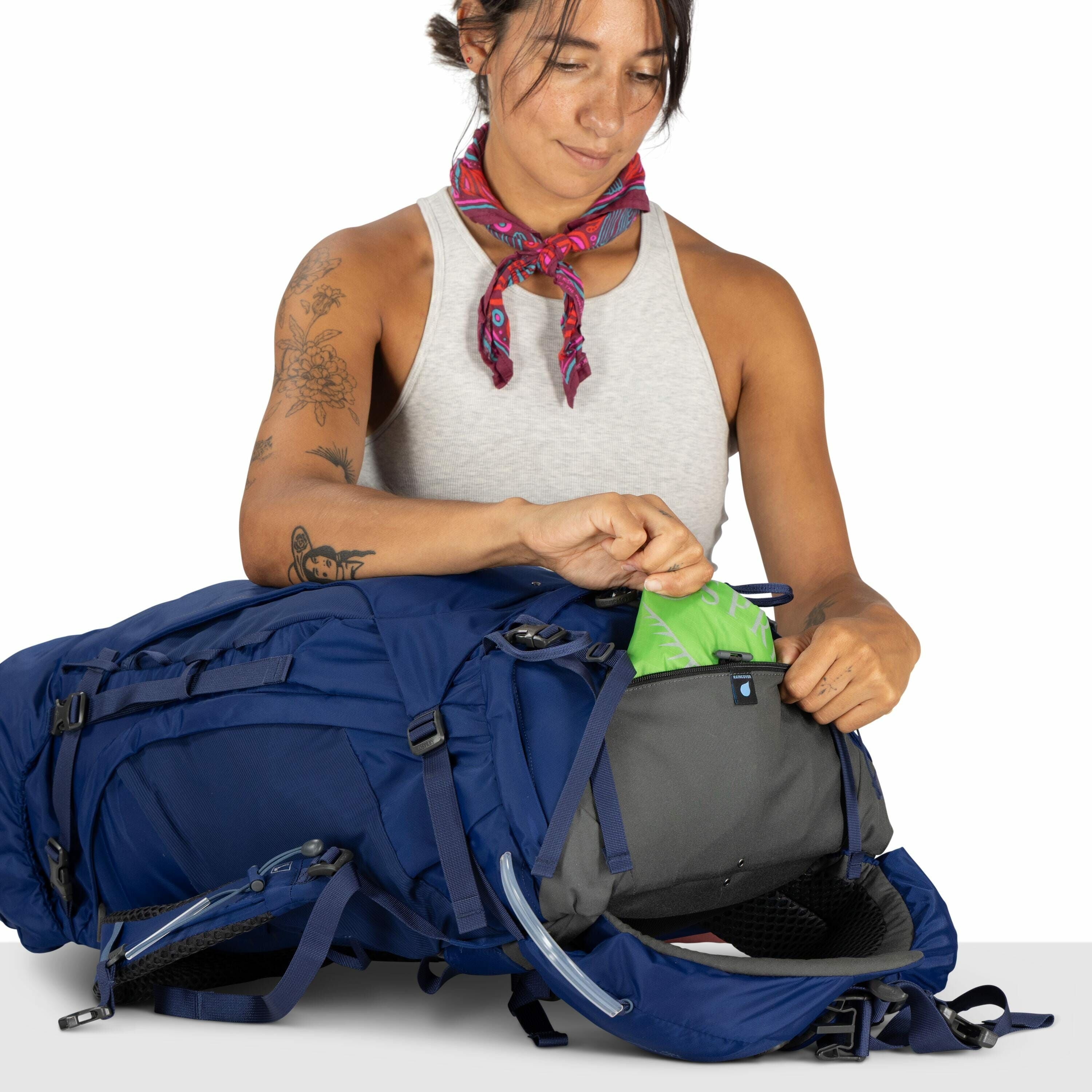 The image shows a person wearing a sleeveless gray shirt and a colorful scarf tied around their neck. They are handling a large blue backpack, which is positioned horizontally on a surface in front of them. The person is seen inserting a bright green item