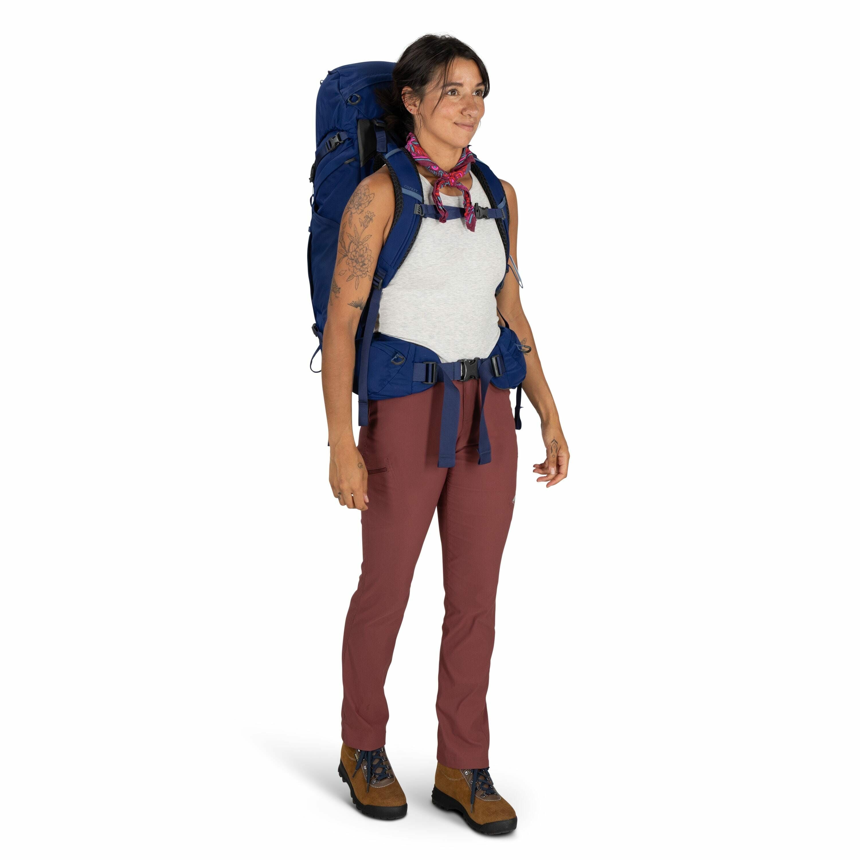 The image depicts an individual dressed for outdoor hiking or backpacking. They are wearing a sleeveless white tank top and reddish-brown pants. The person is equipped with a large blue backpack, which has padded shoulder straps and a belt around the wais