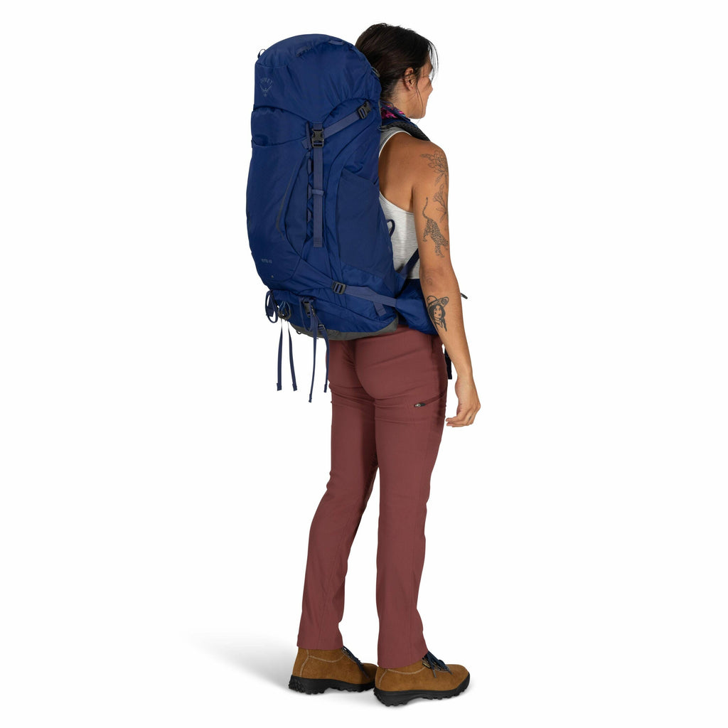 The image depicts a person standing, equipped for hiking. They are wearing a sleeveless gray top and burgundy pants that are slightly form-fitting and have a pocket on the right thigh. They are carrying a large blue backpack with multiple straps and compa