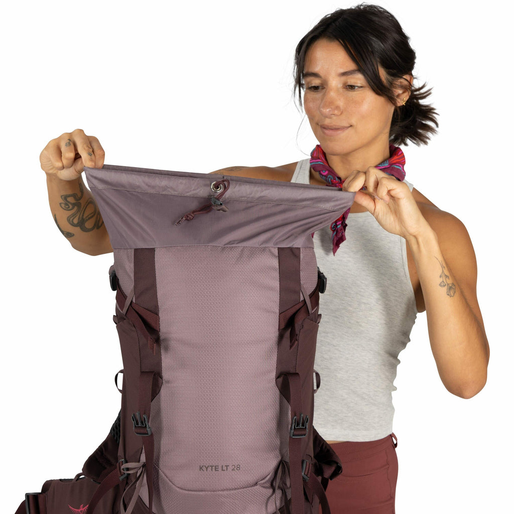 The image features a person holding up the top flap of a backpack, revealing the interior closure mechanism. The backpack is labeled "Kyte LT 28" and appears to be a sturdy outdoor backpack, designed for hiking or trekking. The backpack has a mix of purpl