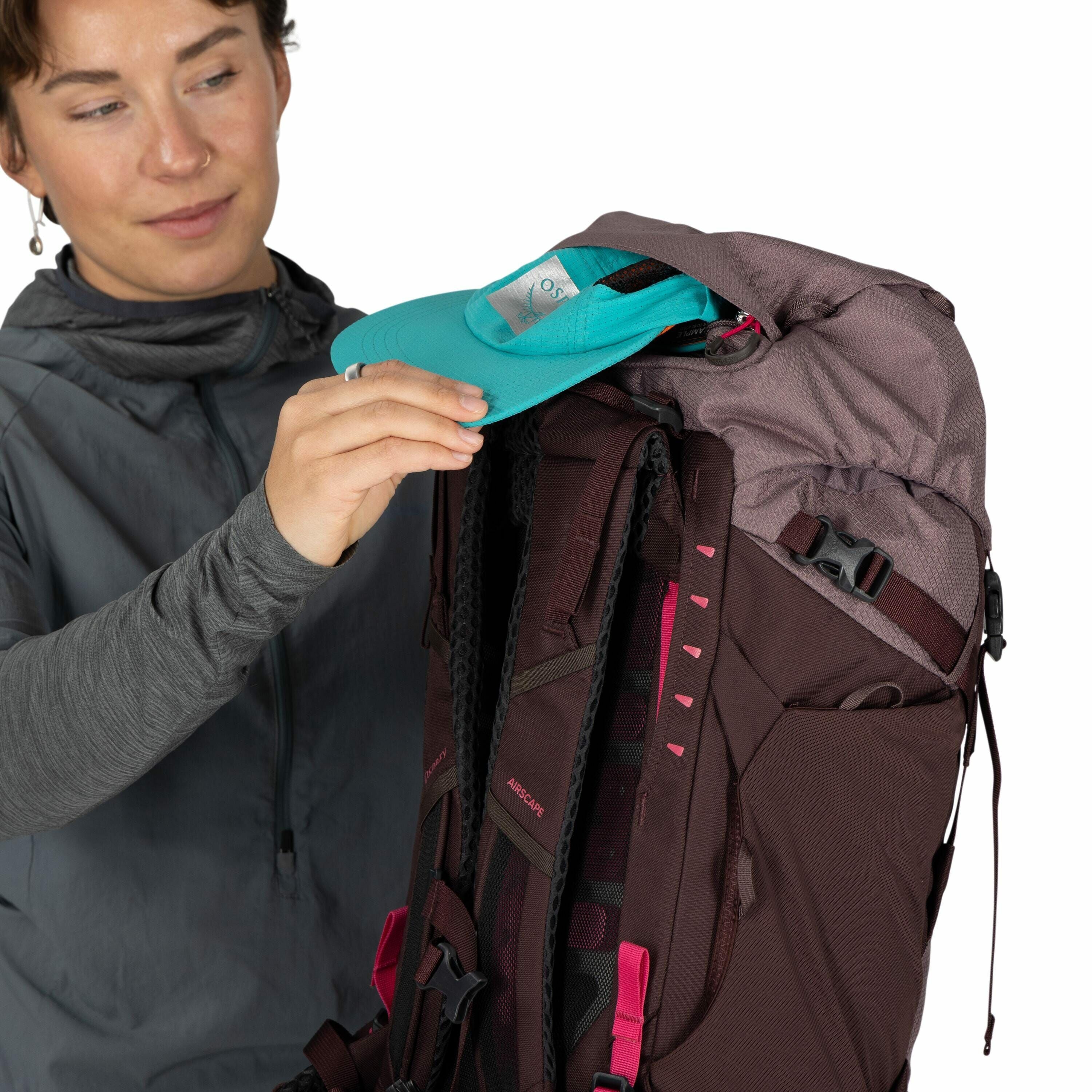 The image shows a person dressed in a gray, long-sleeved hoodie holding a turquoise cap and packing it into a maroon-colored backpack. The backpack features gray and pink accents, with visible straps and buckles. The cap, held by the person, has the "OSPR