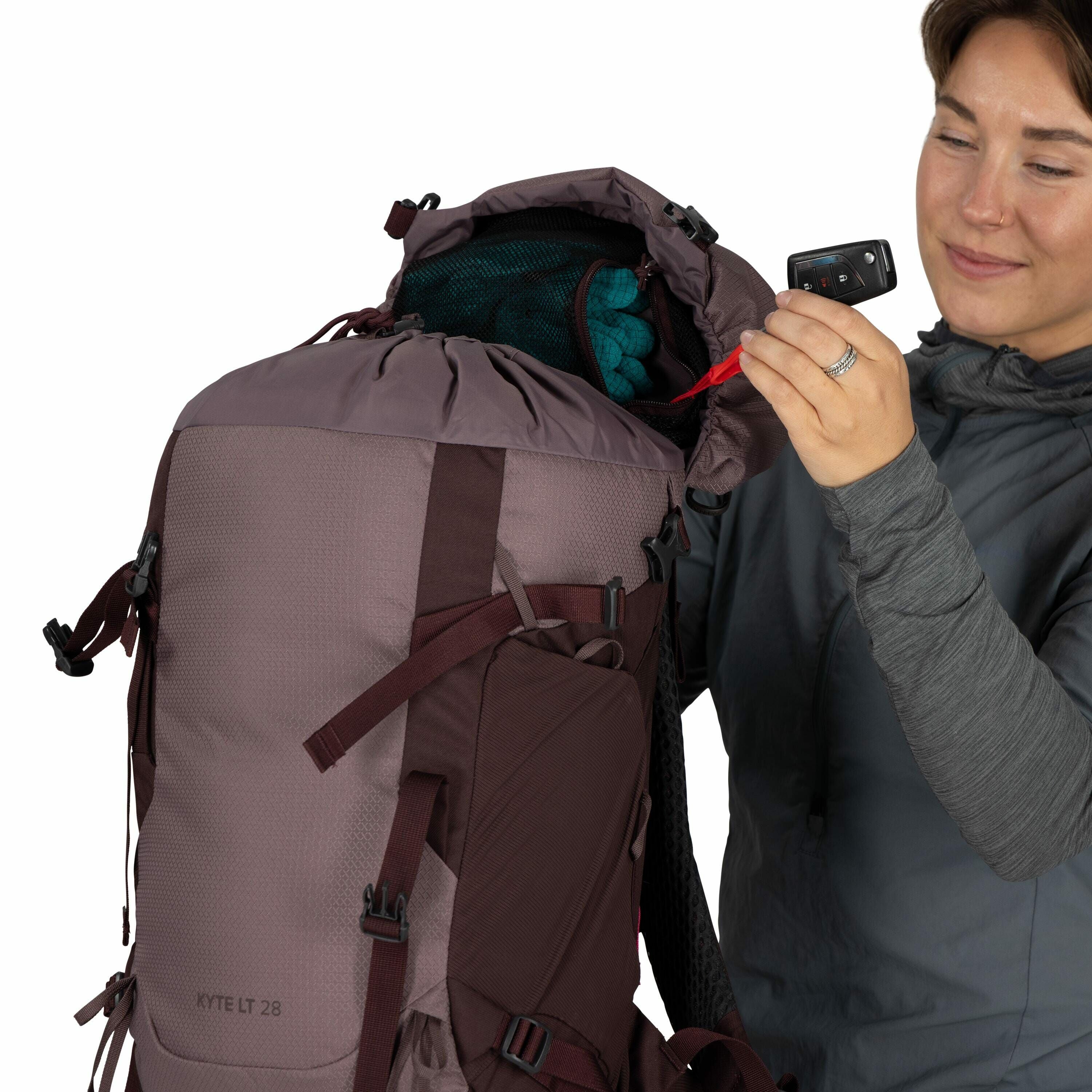 The image shows a person wearing a gray long-sleeved top and holding a maroon and gray backpack with a label "KYTE LT 28" on it. The backpack appears to be open at the top, revealing a teal-colored item, possibly clothing, inside the main compartment. The