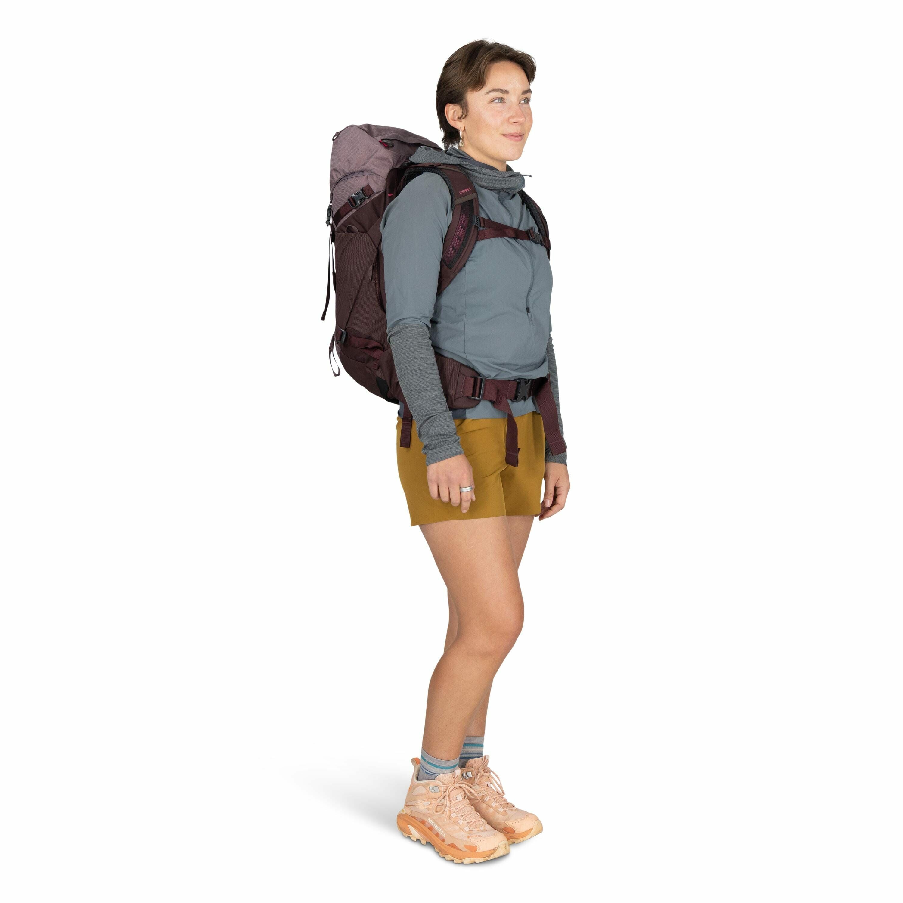 The image shows a person dressed for hiking or trekking, carrying a large backpack with several compartments and straps. The individual is wearing a light blue long-sleeve shirt with thumbholes, paired with brown shorts. They have hiking boots on, which a