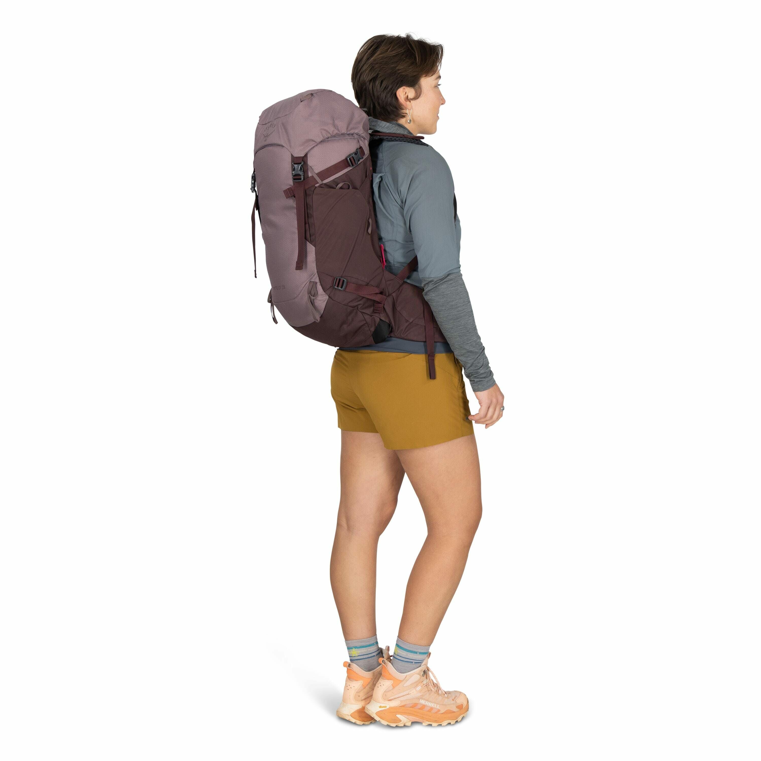 The image features a person standing sideways, showcasing a large backpack worn on their back. The backpack is predominantly maroon with black accents, equipped with multiple straps for adjustments and support. The individual is dressed in outdoor attire,