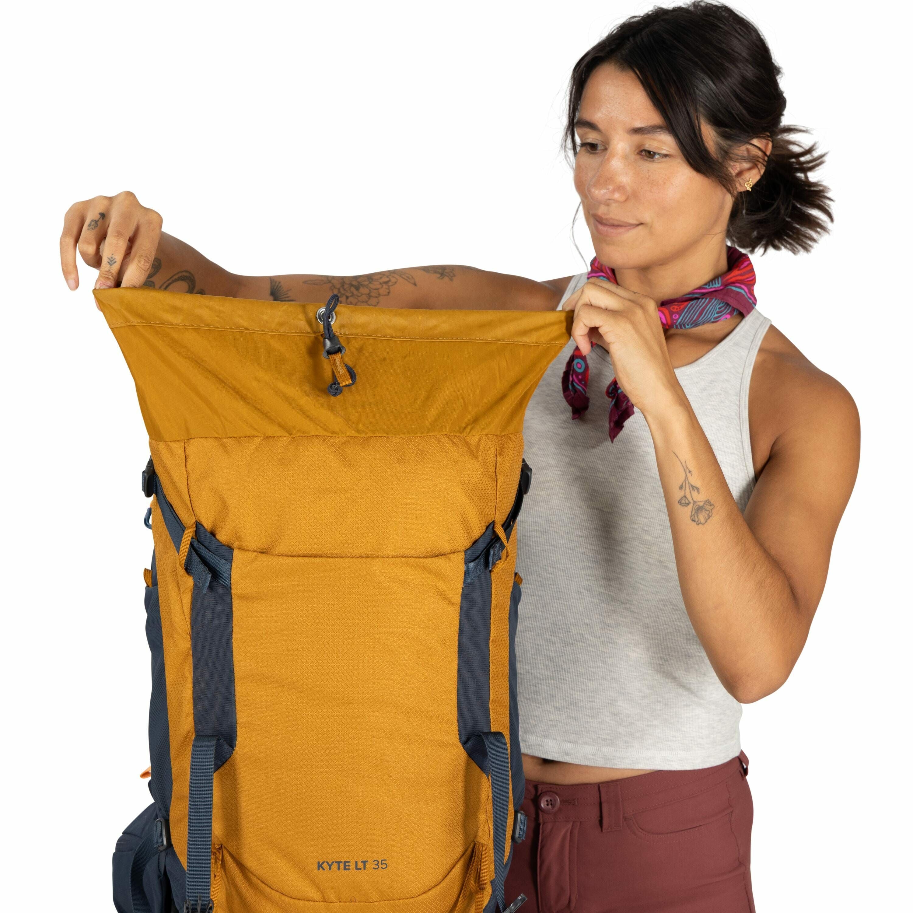 The image depicts a person wearing a gray sleeveless top and reddish-brown pants, holding an orange backpack labeled "KYTE LT 35." The backpack features blue straps and multiple compartments, designed for practical use and outdoor activities. The person h