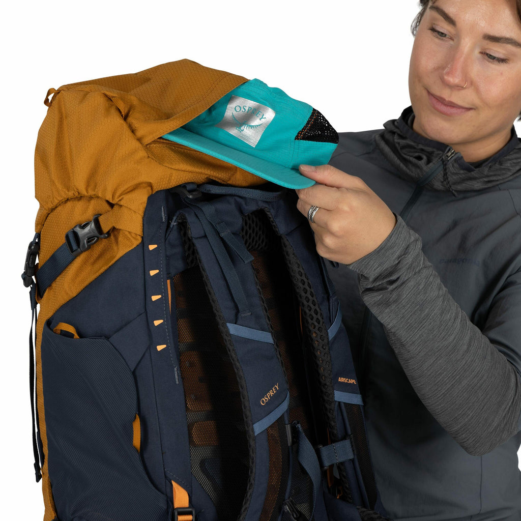In the image, a person dressed in a gray outdoor jacket is holding an Osprey backpack with one hand, while the other hand is lifting the top flap. The backpack features a combination of mustard yellow and navy blue colors, with visible labels on the back