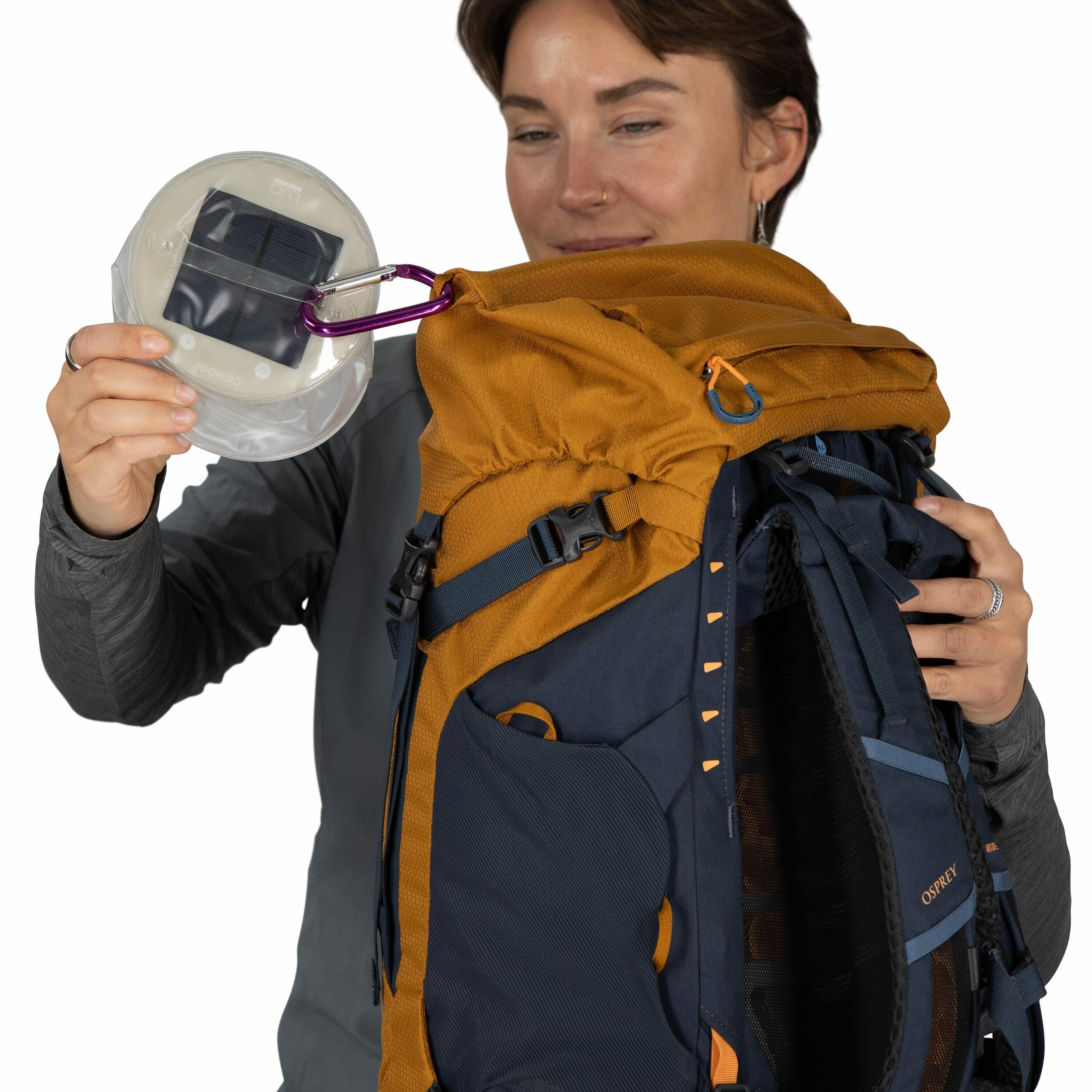 The image shows a person holding an Osprey backpack, which has a prominent mustard yellow and dark blue color scheme. The person is also holding a small circular solar-powered device, possibly a lantern, which is attached to the backpack with a purple car