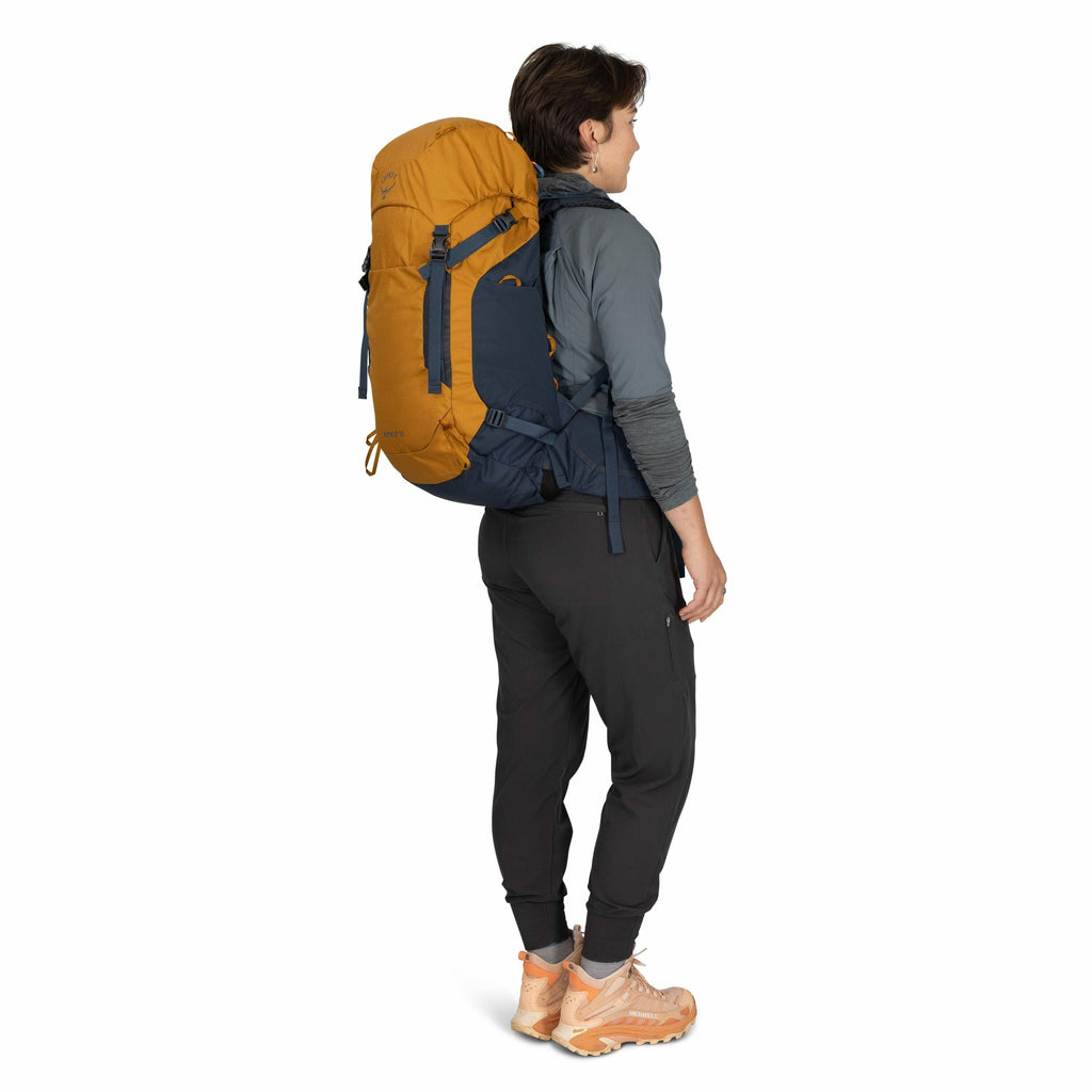 The image shows a person standing sideways, wearing outdoor clothing suitable for hiking or trekking. They are equipped with a large yellow and dark blue backpack labeled "ATLAS," which features multiple straps and adjustments for secure fitting. The pers