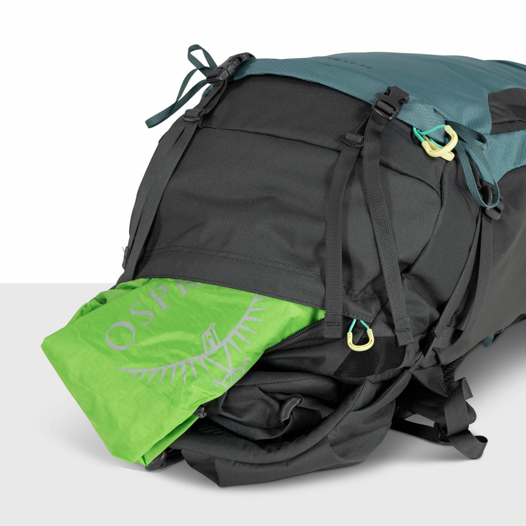 The image shows a close-up of a black and teal backpack that has a bright green item, with the Osprey logo visible, partially tucked inside a pocket. The backpack is designed with multiple adjustable straps and clips, including two visible black buckles a
