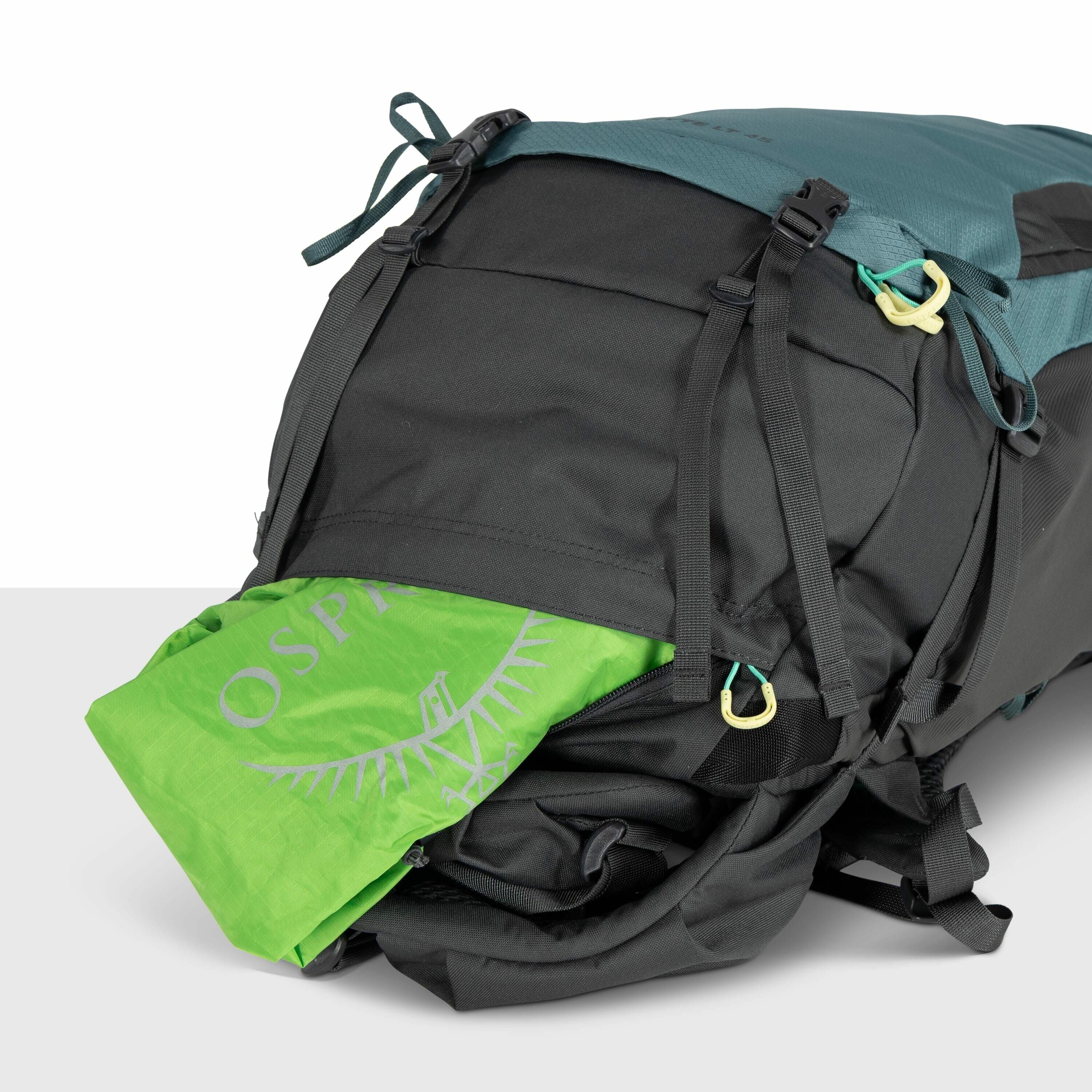 The image shows a close-up of a black and teal backpack that has a bright green item, with the Osprey logo visible, partially tucked inside a pocket. The backpack is designed with multiple adjustable straps and clips, including two visible black buckles a