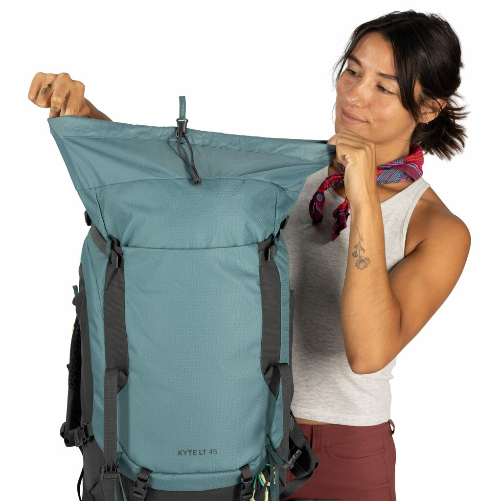 The image features a person holding up an Osprey Kyte LT 45 backpack. The backpack is a shade of blue-green with black straps and accents. It appears to be designed for outdoor activities like hiking or backpacking, as indicated by its large size and stur