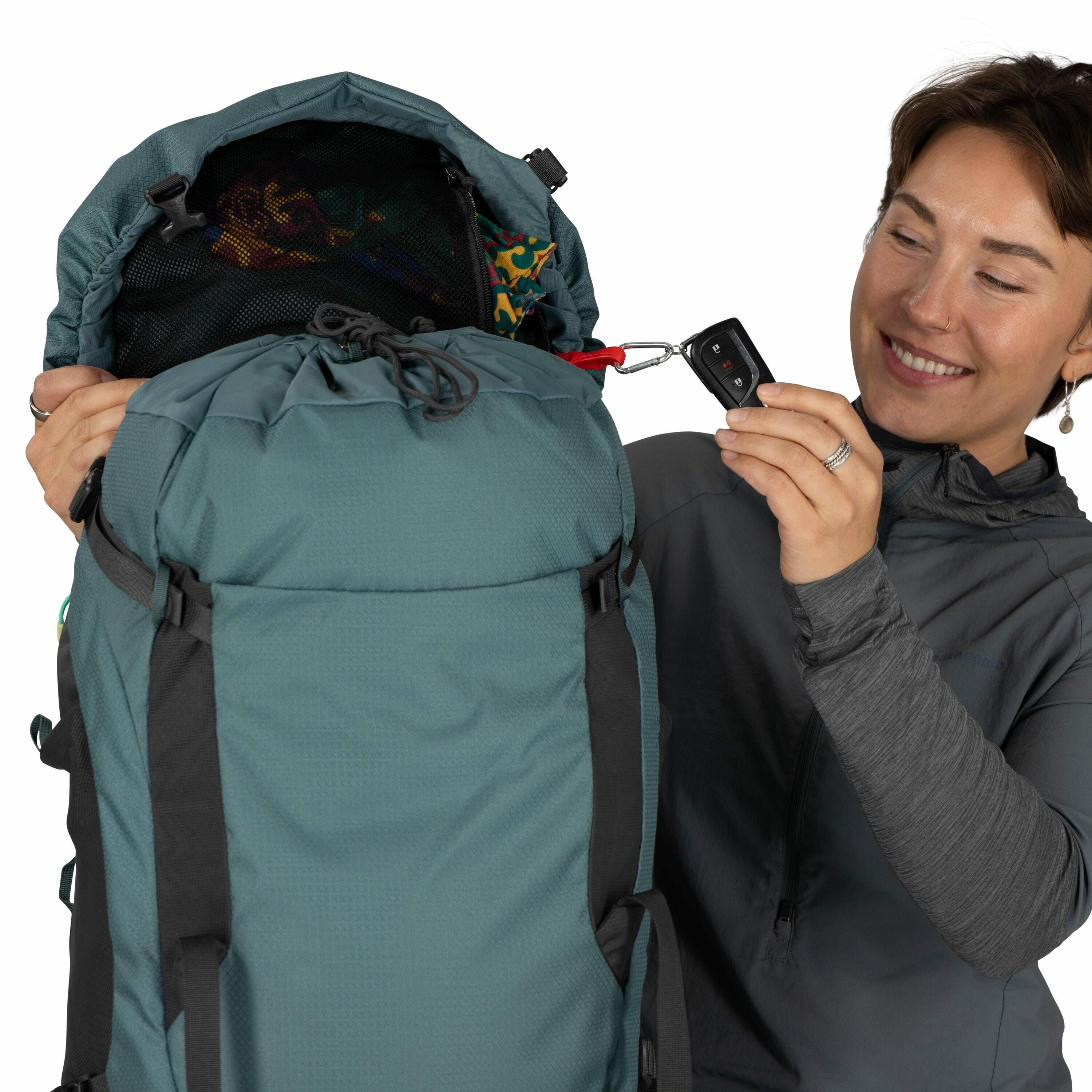 The image shows a person holding a large teal-colored backpack. The backpack has a top flap that is open, revealing some colorful fabric inside. The person is dressed in a gray long-sleeve shirt and appears to be demonstrating the use of a carabiner attac