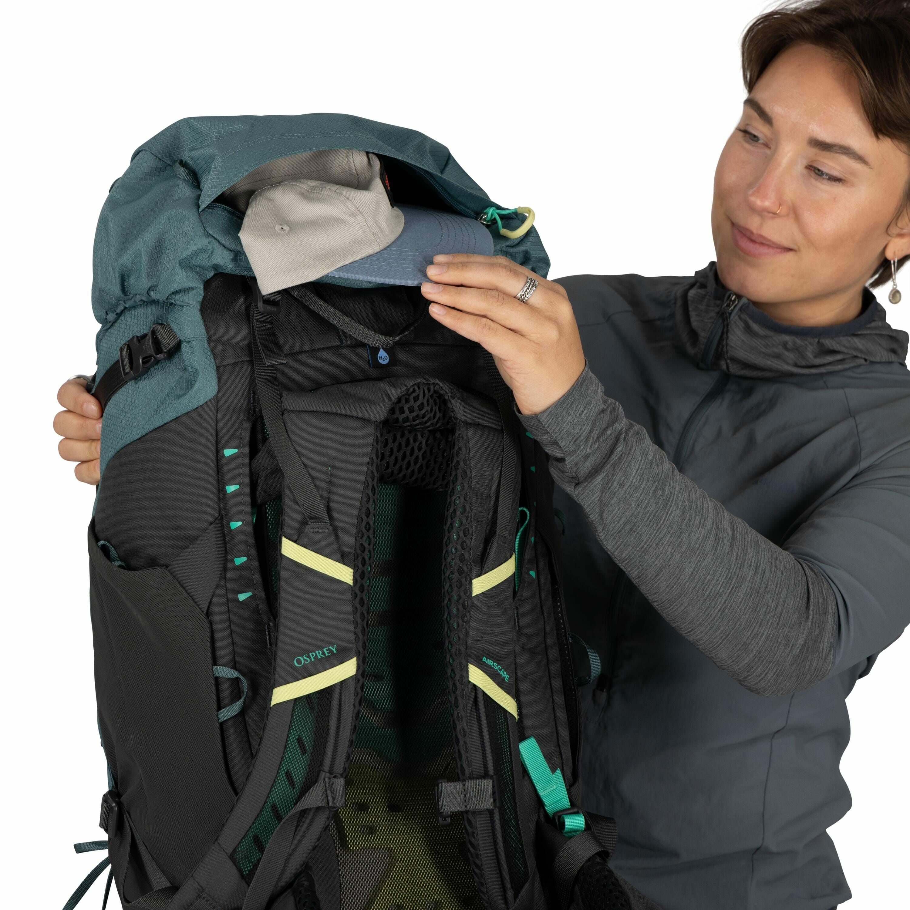 The image features a person holding an Osprey backpack, which is predominantly black with teal accents. The individual is dressed in a gray long-sleeved top and appears to be demonstrating the backpack's features by opening a top compartment to reveal a l