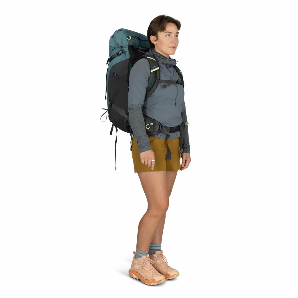 The image shows a person dressed in outdoor hiking attire, equipped for a trek. They are wearing a large, fully packed backpack that is secured with multiple straps, featuring a color scheme of dark gray and teal. The individual is dressed in a blue-gray