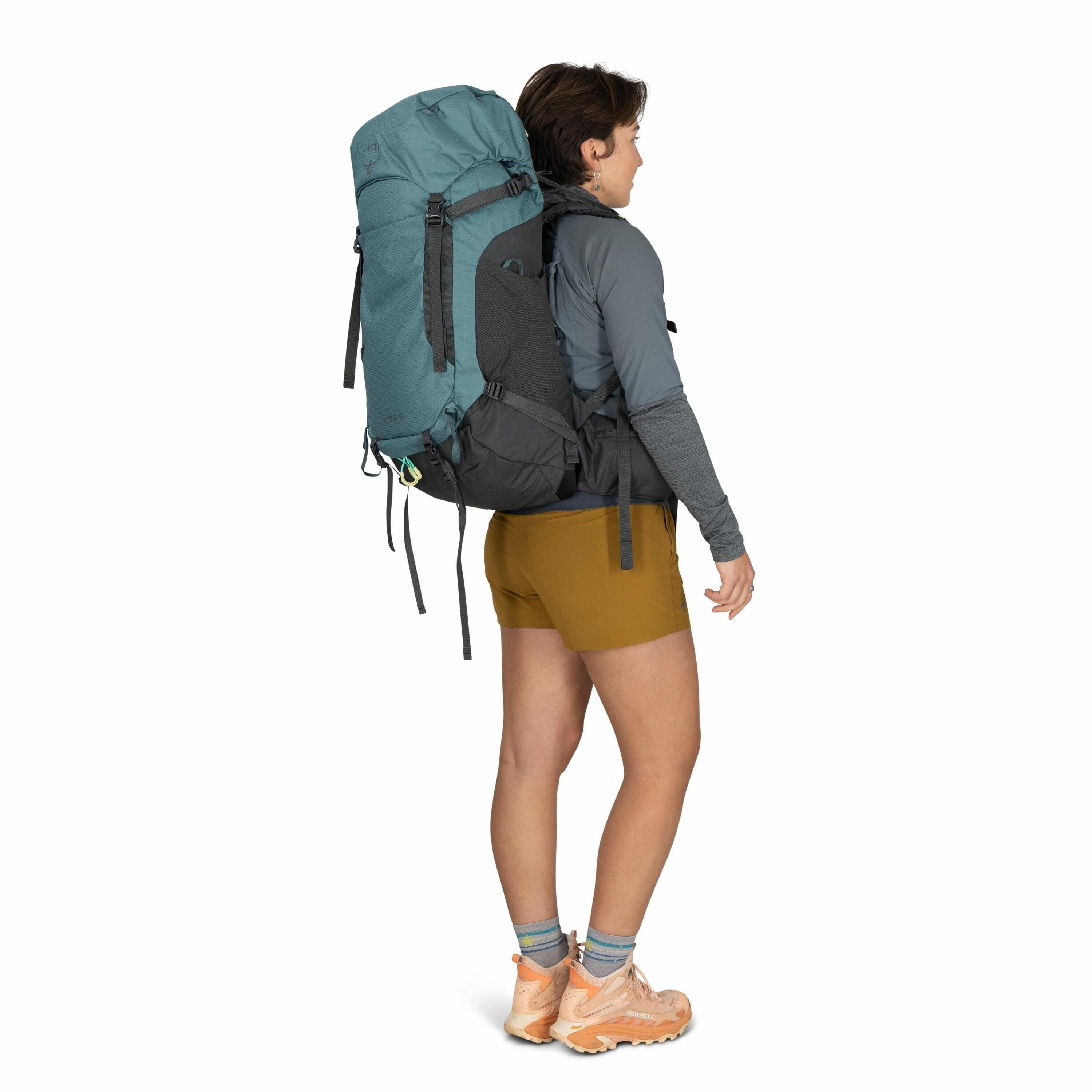 The image shows a person dressed in outdoor hiking attire. They are wearing a long-sleeved, dark gray shirt paired with mustard-colored shorts. On their back, they are carrying a large, teal and black hiking backpack secured with multiple straps. Their fe