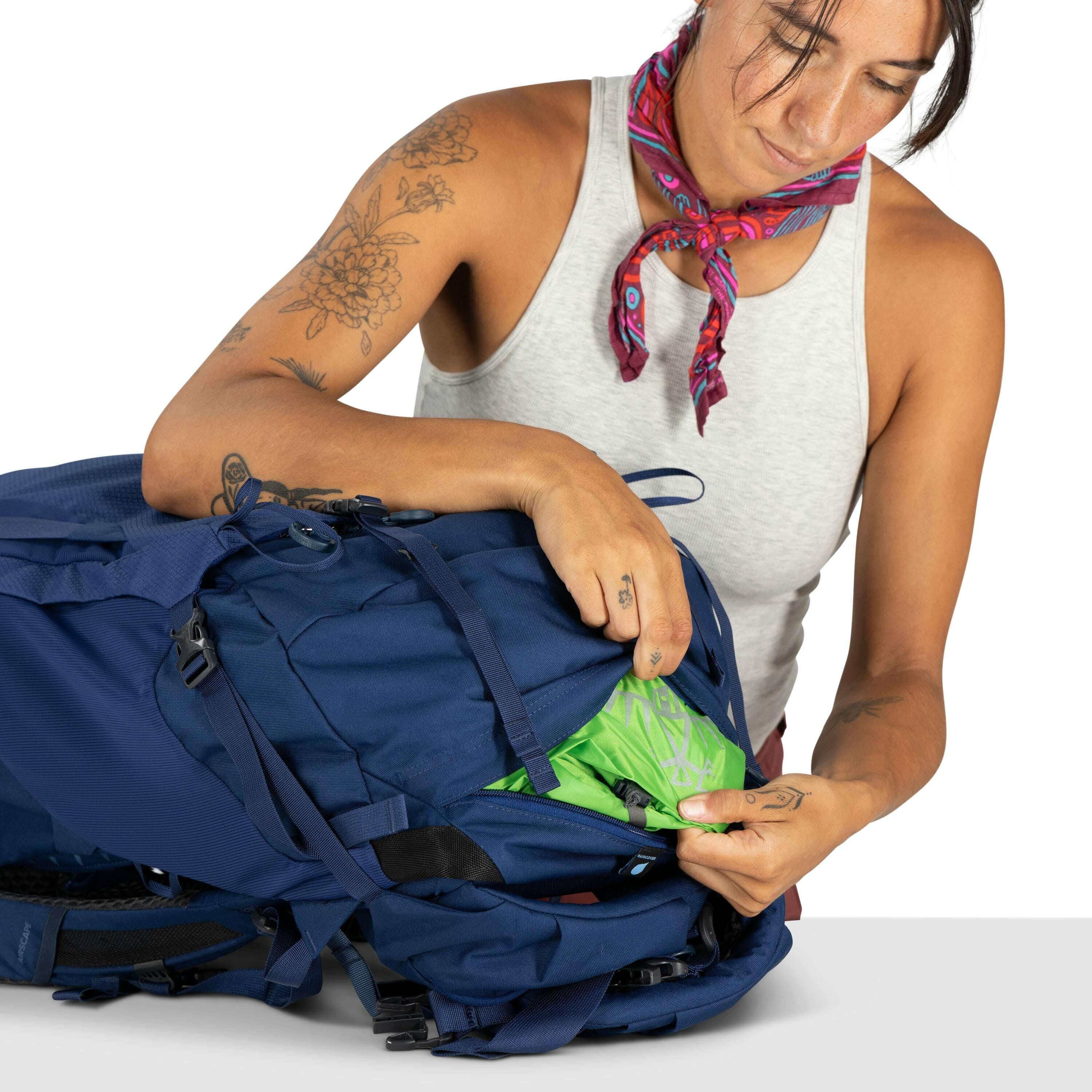 The image shows a person interacting with a large blue backpack. The individual is wearing a sleeveless gray top, displaying several floral tattoos on their arms, as well as a red scarf tied around their neck. They are in the process of accessing a zipper