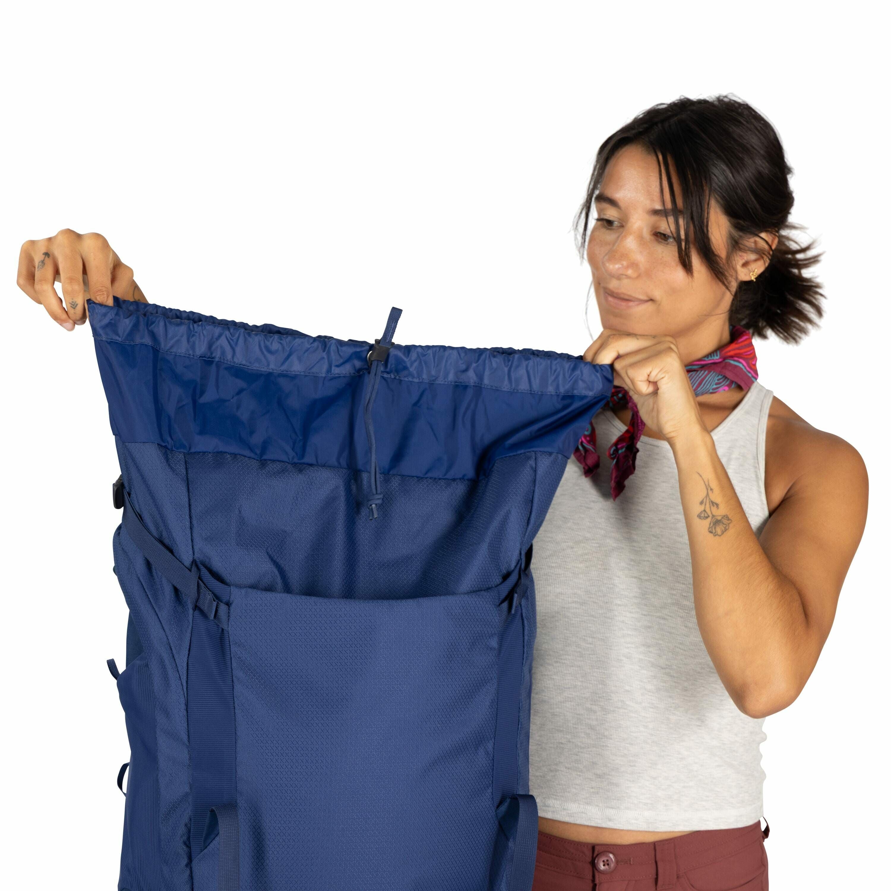 The image features a person holding a large blue backpack with both hands, showcasing its open top section. The person is dressed in a casual grey sleeveless top and rust-colored pants, accessorized with a colorful scarf around their neck. A small floral