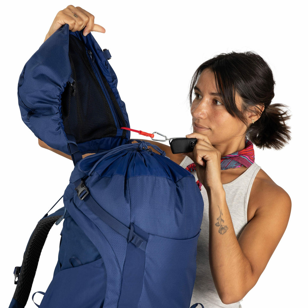 The image shows a person demonstrating the functionality of a blue backpack. The backpack has a large main compartment with a lid that includes a zippered pocket. The individual is using a red strap with a buckle attached to the backpack to show how it ca