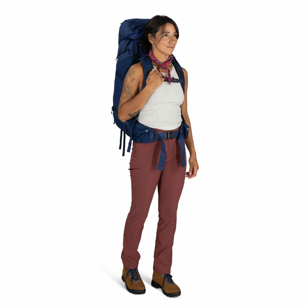 The image shows a person dressed in outdoor gear, ready for a hiking or backpacking adventure. They are wearing a sleeveless gray top, brick-colored pants, and sturdy brown and blue hiking boots. A large blue backpack is secured on their back with padded