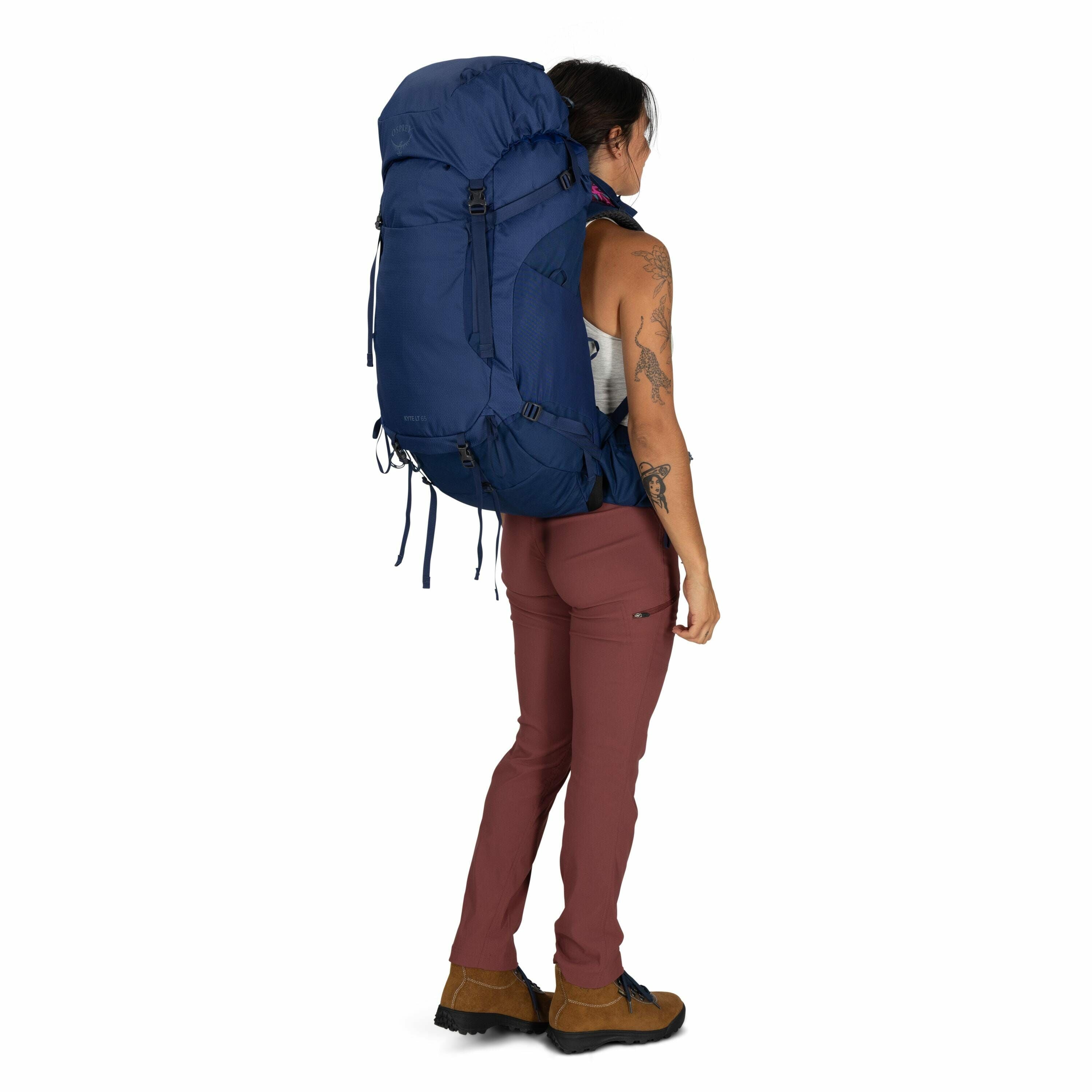 The image shows a person from a side view standing with a large, blue backpack strapped onto their back. They are wearing a white tank top and pinkish-brown pants, along with tan hiking boots. The backpack appears to be designed for hiking or backpacking,