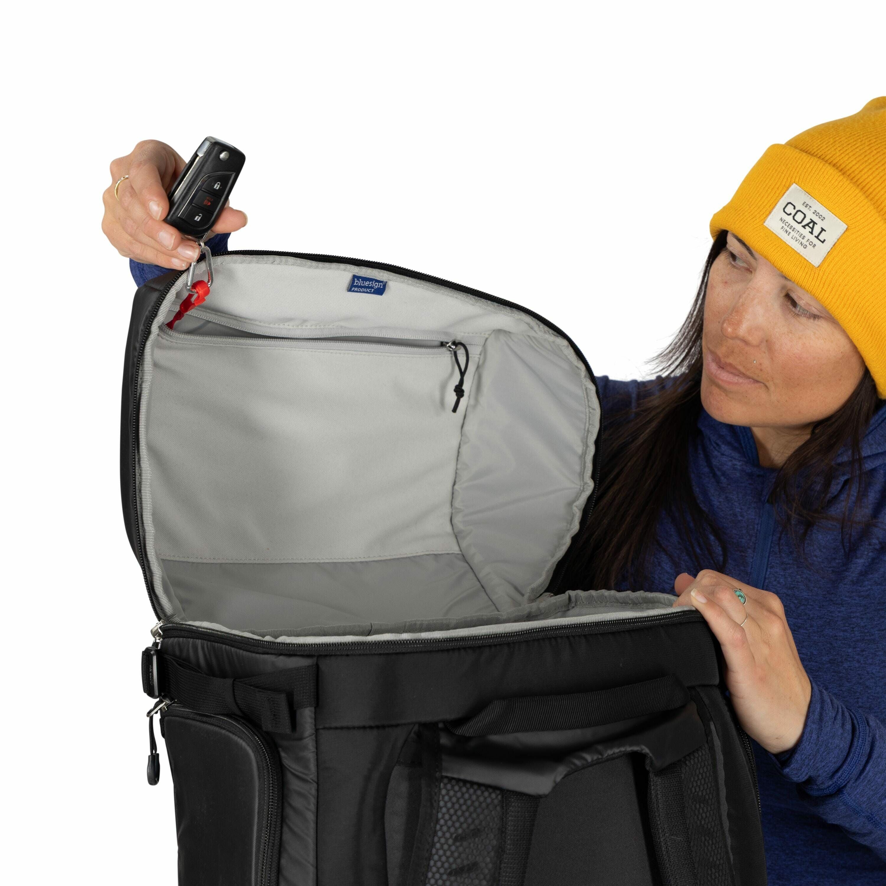 The image shows a person wearing a bright yellow beanie and a blue long-sleeve shirt, holding a black bag open. The interior of the bag is grey, and the person is pulling the bag's lid upwards to reveal its spacious compartment. In the person's left hand,