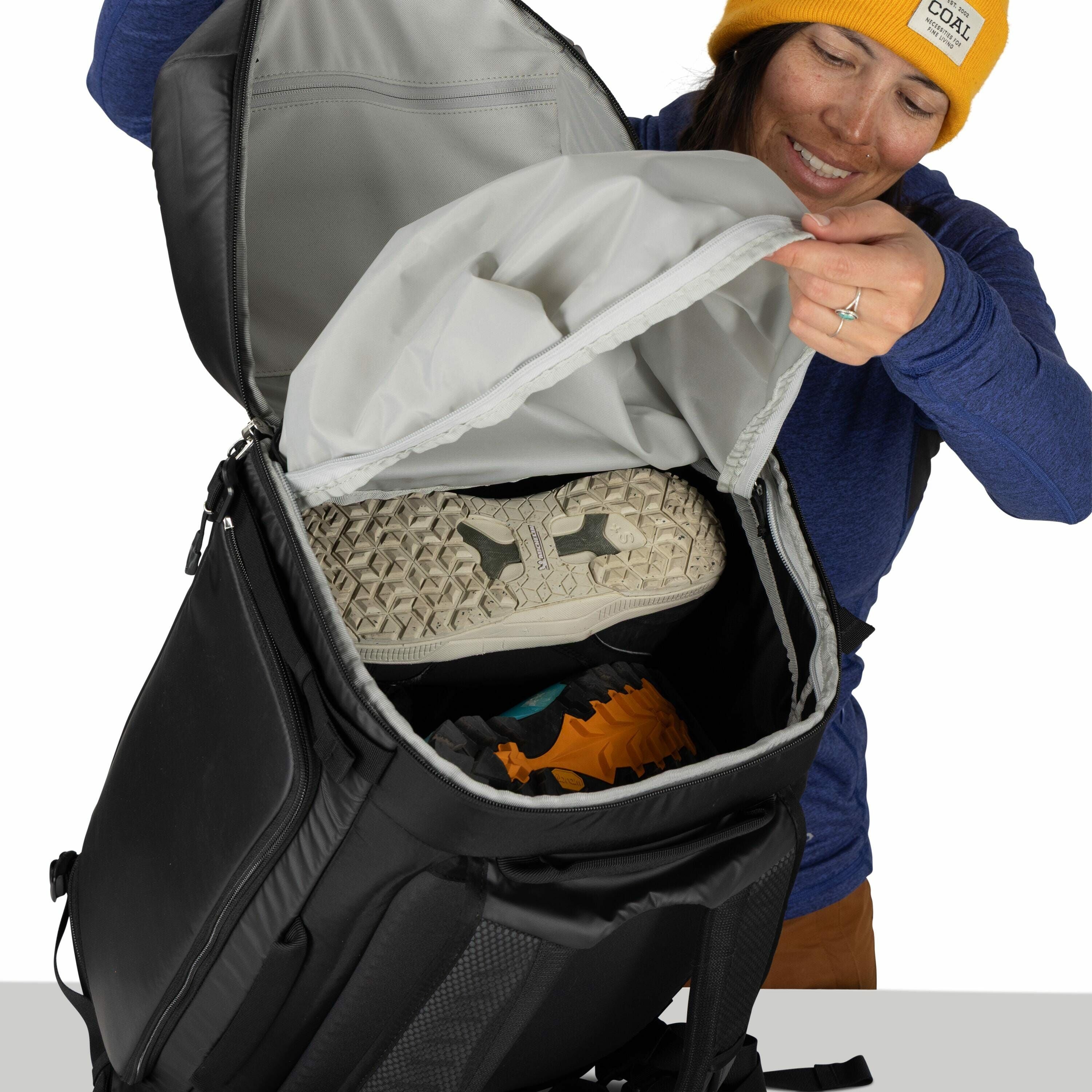 The image shows a person wearing a blue long-sleeve shirt and a yellow hat, holding open the flap of a black backpack to reveal its contents. Inside the backpack, there is a neatly packed pair of shoes with a unique, textured sole visible at the top. Belo