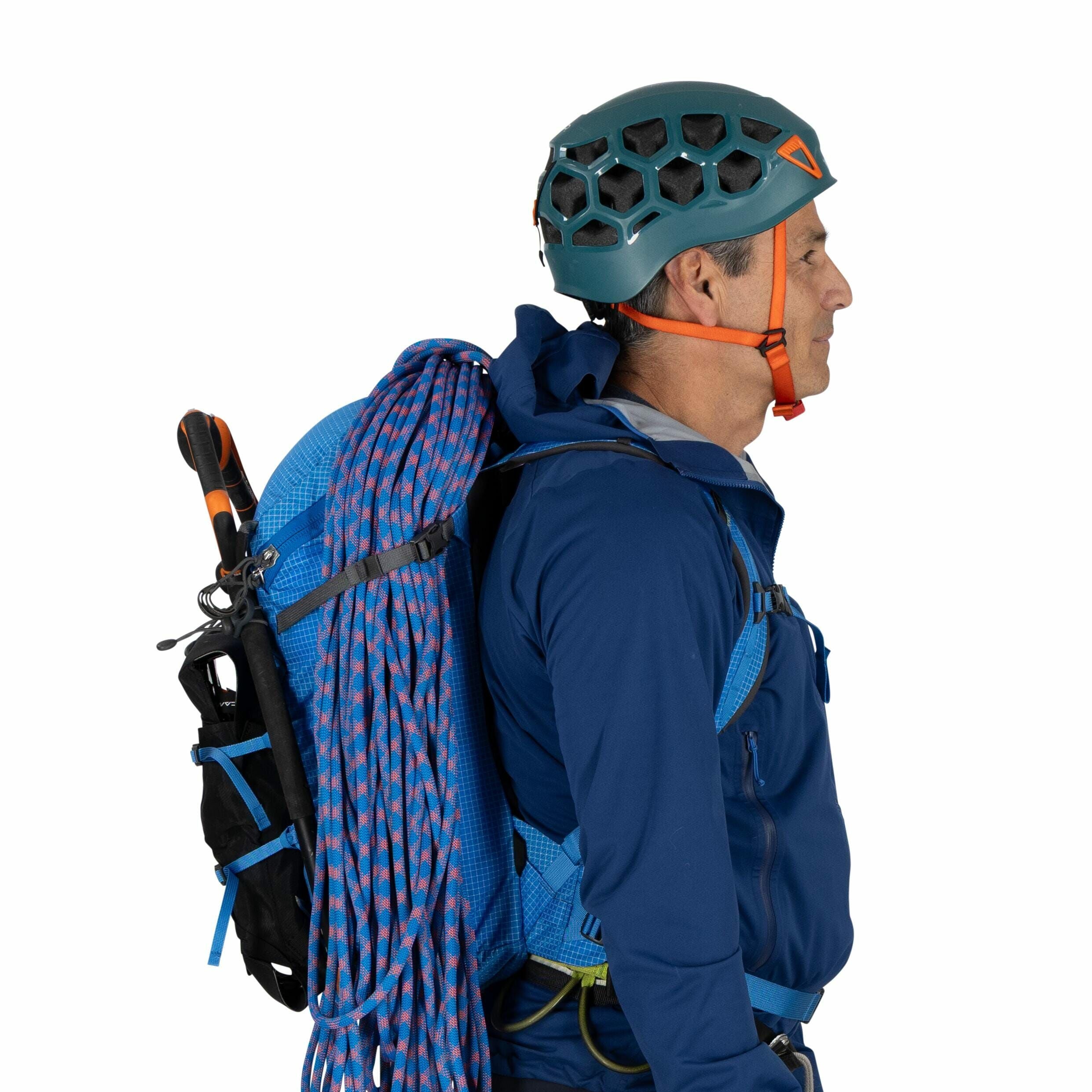 The image shows a person dressed in outdoor climbing gear, viewed from the side. They are wearing a teal climbing helmet with an open hexagonal pattern and orange accents, along with a blue jacket and backpack. The backpack is equipped with climbing essen