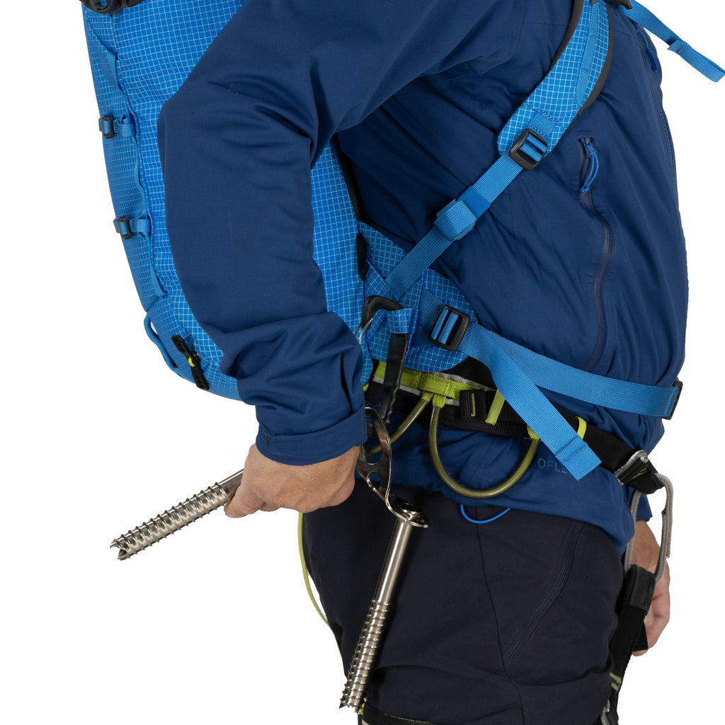 The image shows a person wearing outdoor gear, likely for climbing or hiking, featuring a blue backpack with multiple straps for secure fitting. The individual is dressed in a matching blue jacket and navy blue pants, indicating a coordinated outfit suita