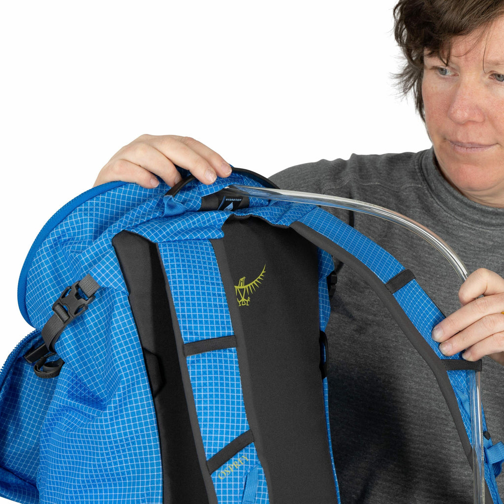 The image shows a person demonstrating a blue backpack, which has a distinctive checkered pattern and is equipped with black padded shoulder straps. A hydration tube runs over one of the shoulder straps, suggesting the backpack has a hydration reservoir o