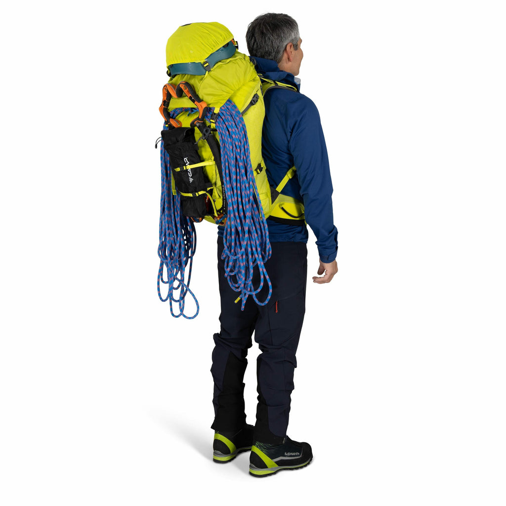 The image shows a man dressed in outdoor hiking or climbing gear, standing and facing away from the camera. He is wearing a blue jacket and dark pants, along with boots that appear to be suitable for hiking or mountaineering. On his back, he carries a lar