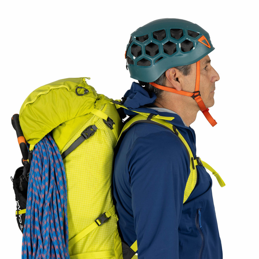 The image shows a person dressed for outdoor activities, likely rock climbing or mountaineering. They are wearing a blue jacket and a bright green climbing backpack. Attached to the backpack is a coiled climbing rope with a blue and red pattern. The perso