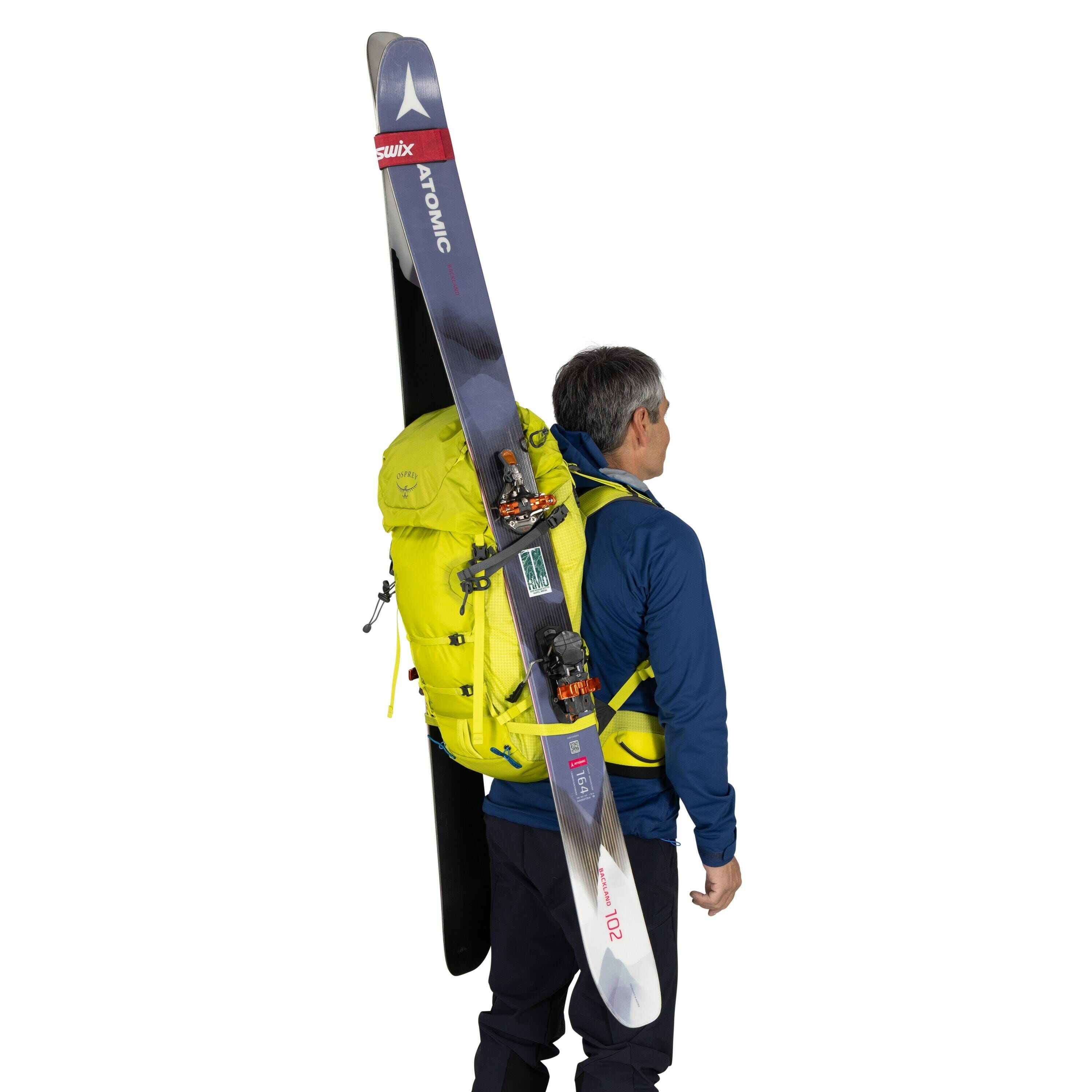 The image features a man standing with his back to the camera, wearing a vibrant yellow backpack that securely holds a pair of skis. The skis, branded "Atomic," have a sleek design with a predominantly dark color scheme and hints of red and white. The man