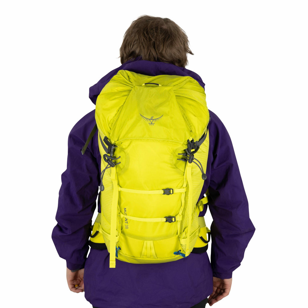 The image shows a person wearing a bright yellow backpack, which appears to be designed for hiking or outdoor activities. The backpack has multiple straps and buckles for securing items and ensuring a snug fit. The person is wearing a purple jacket, which