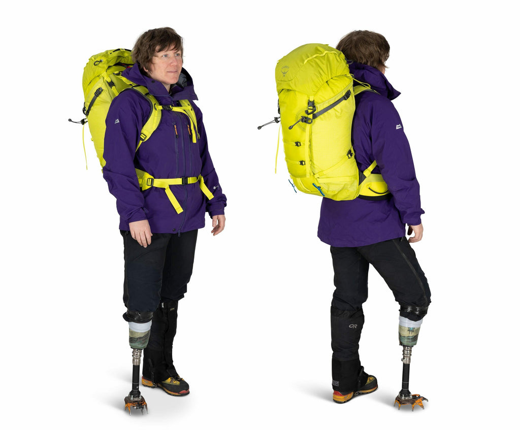 The image shows a person dressed in outdoor hiking gear, featuring a purple jacket and black pants. They are carrying a bright yellow backpack equipped with various straps and compartments, suitable for hiking or trekking. The individual is also wearing s