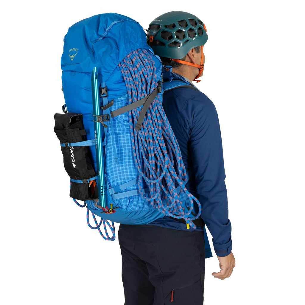 The image shows a person viewed from behind, equipped for a climbing or hiking excursion. They are wearing a blue long-sleeve top and dark pants, with a helmet on their head. The large blue backpack they carry is fully loaded, featuring a coiled climbing