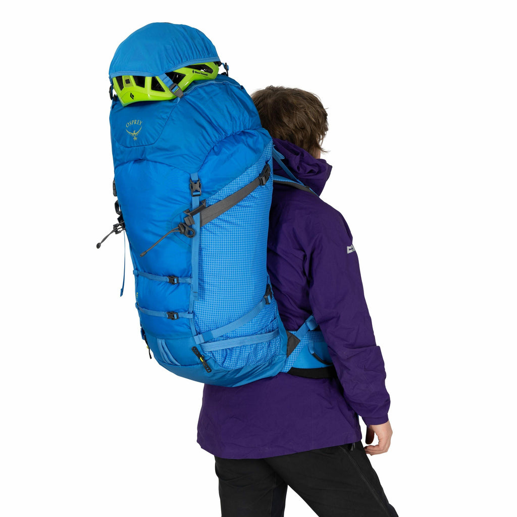 The image shows a person from behind, wearing outdoor gear and carrying a large backpack. The backpack is vibrant blue with gray accents and appears to be designed for hiking or mountaineering, with multiple straps and compartments. On top of the backpack