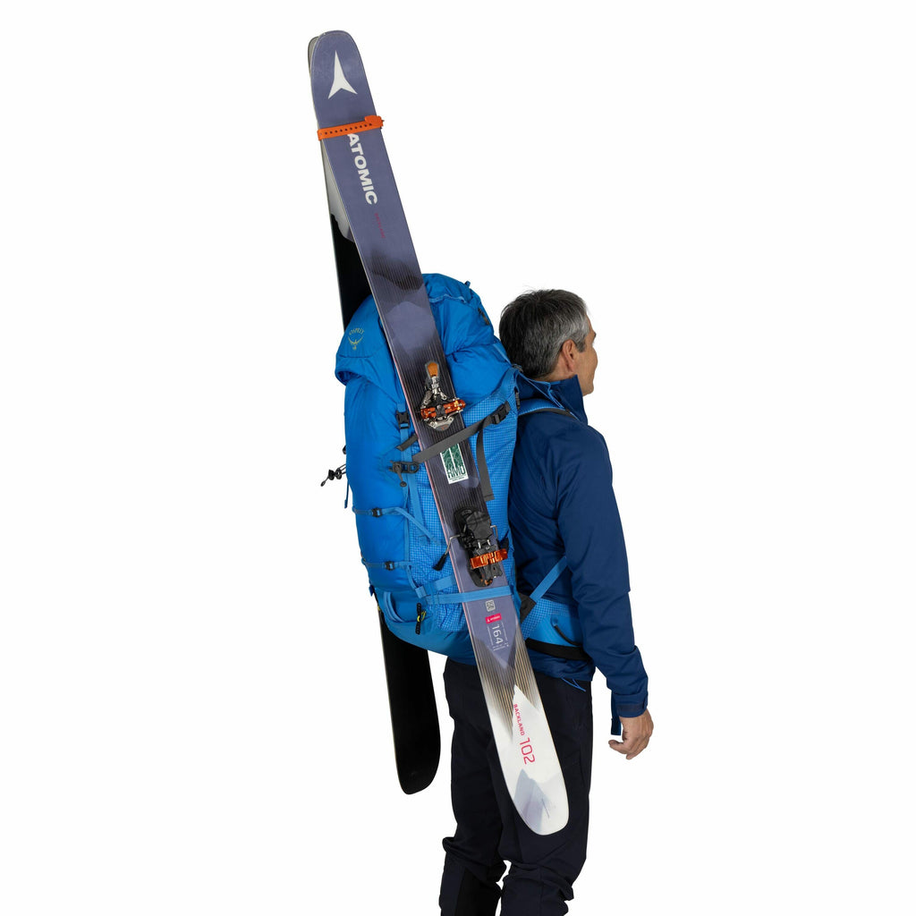 The image shows a person standing sideways, carrying a large blue backpack equipped for winter sports. Attached to the backpack is a pair of skis, with the brand name "Atomic" visible on one of them. The individual is dressed in a blue jacket and dark pan