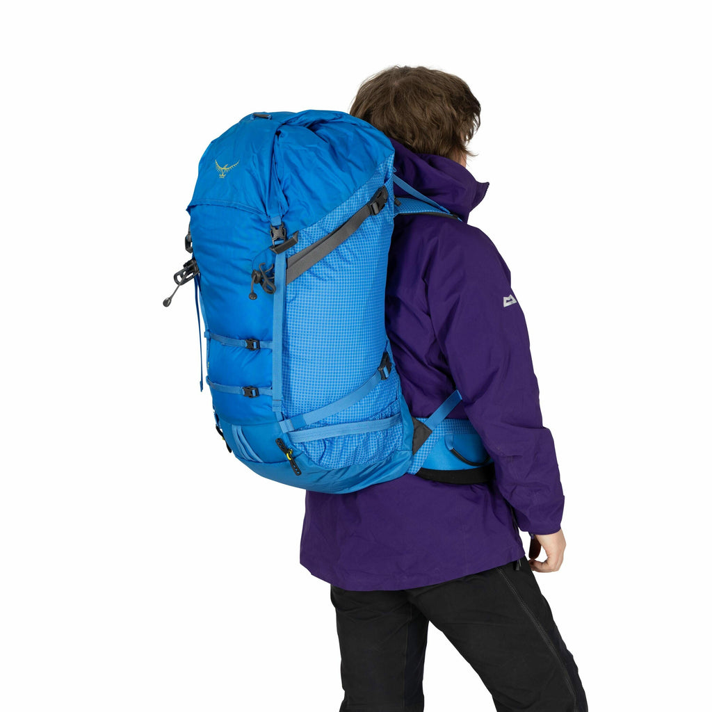 The image shows a person wearing a large blue backpack, designed for outdoor or hiking activities. The backpack is spacious and has multiple straps and compartments, suggesting it's suitable for carrying substantial gear. The person is also wearing a purp