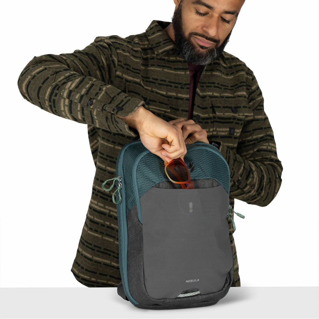 The image shows a person dressed in a patterned, long-sleeve shirt with shades of green and dark accents. They are holding a gray and teal backpack with multiple compartments. The person is in the process of placing or removing a pair of red-framed sungla