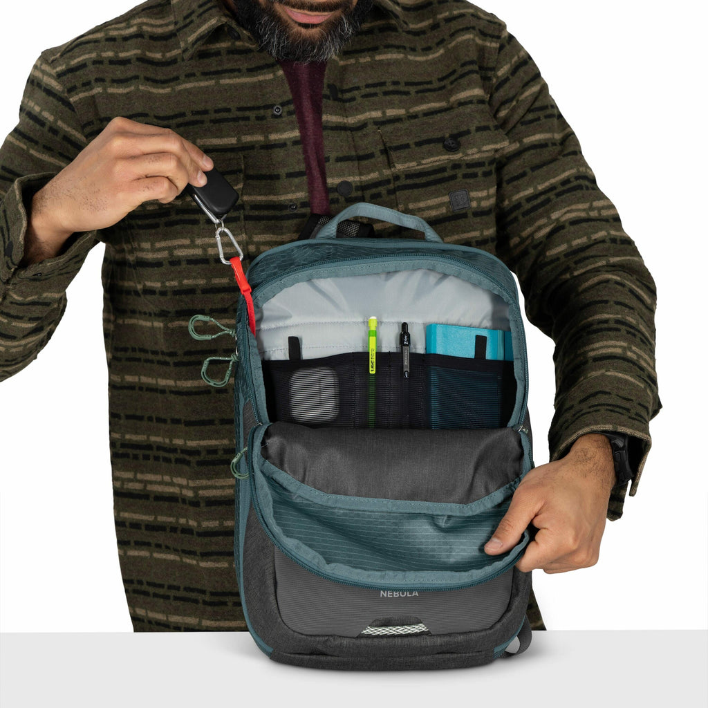 The image shows a person organizing the contents of an open backpack labeled "NEBULA." The backpack is predominantly blue-gray with a front pocket that is partially unzipped, revealing a well-organized interior compartment. The person is attaching or deta