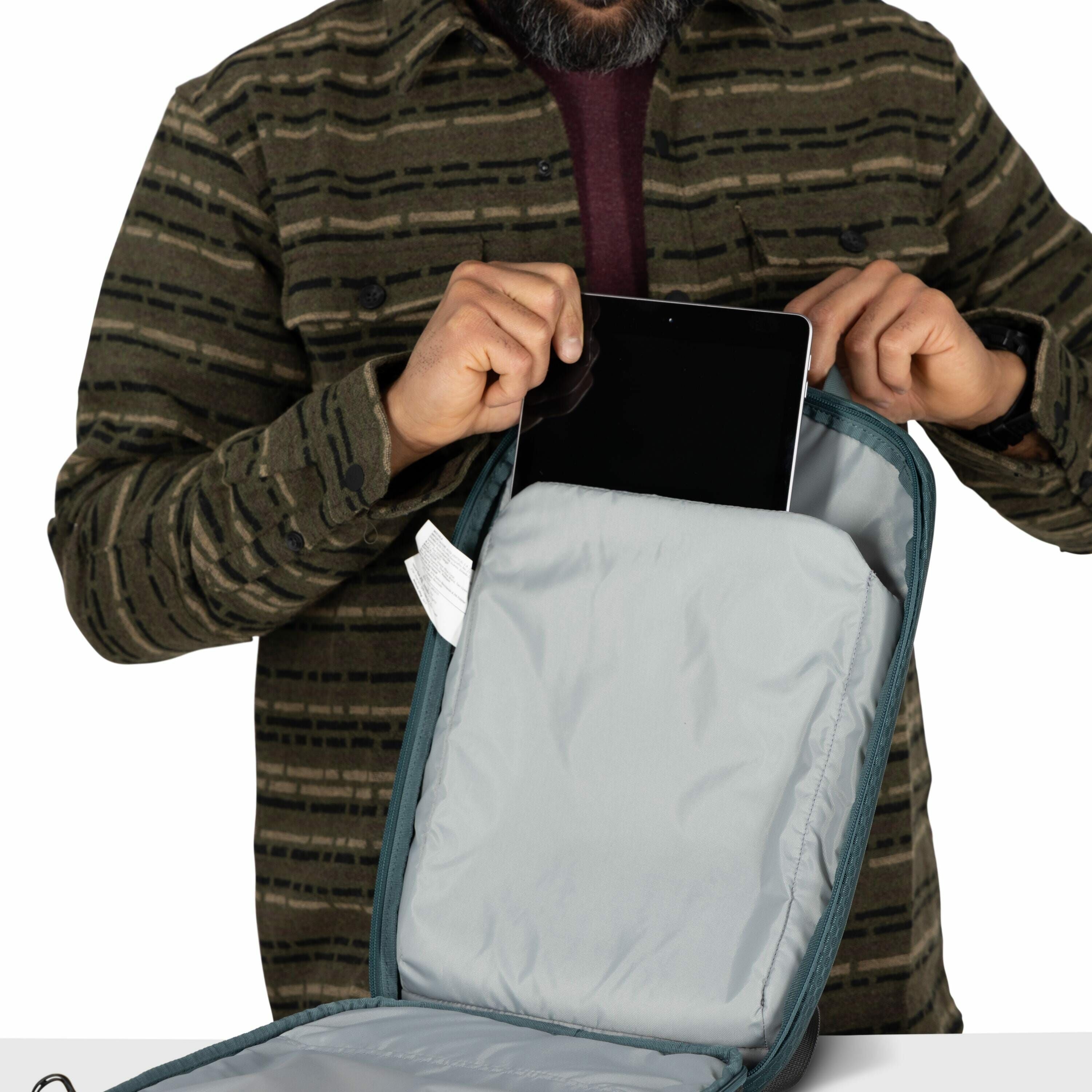 The image shows a man inserting or removing a tablet from a backpack. He is wearing a green and brown striped flannel shirt and has a beard, though his face is not visible in the image. The backpack is opened, revealing a light gray interior where the tab