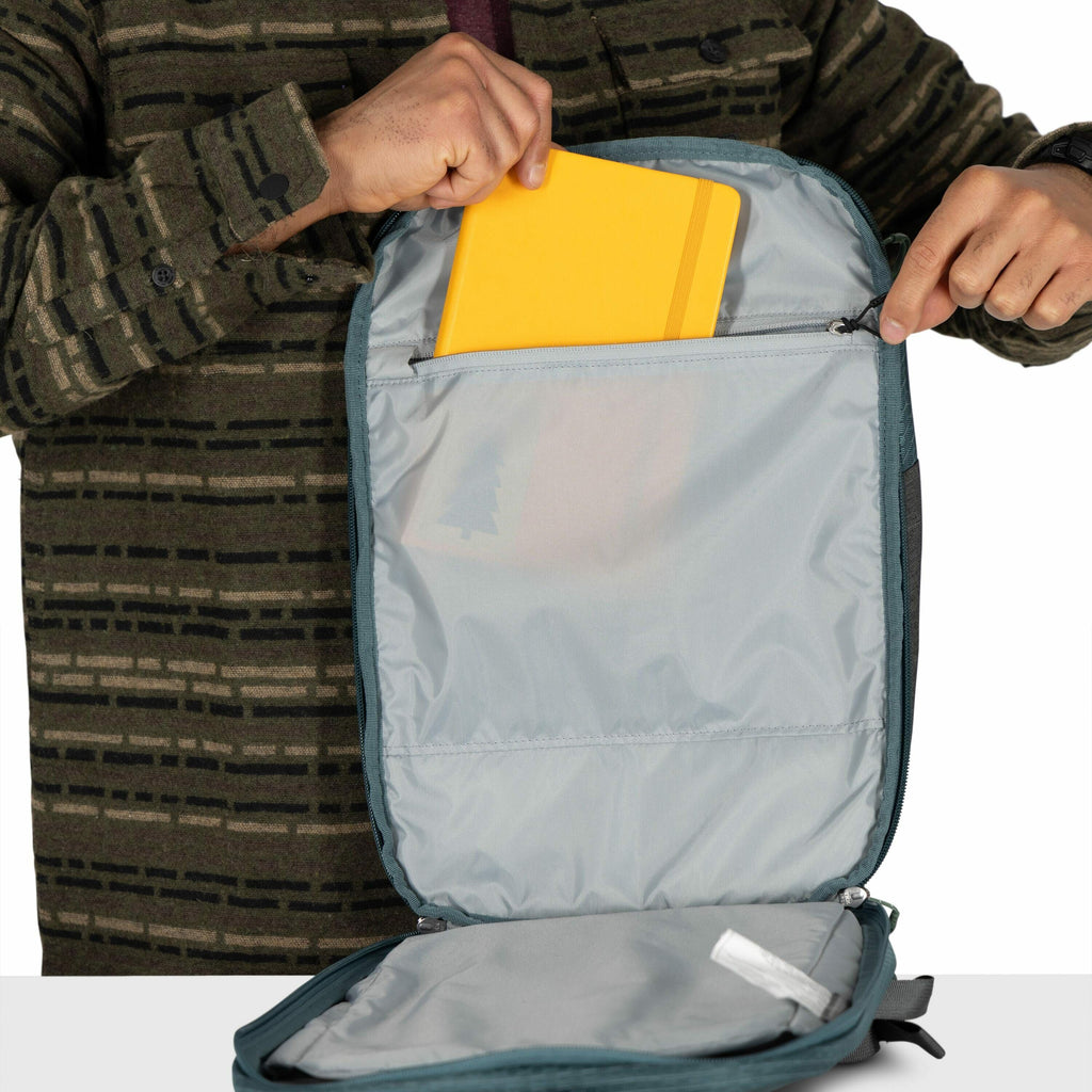 The image shows a person wearing a patterned green jacket, opening a blue-grey backpack. The backpack is open, revealing an inner compartment with a zipper, and the person is placing or removing a bright yellow folder or tablet from this compartment. The