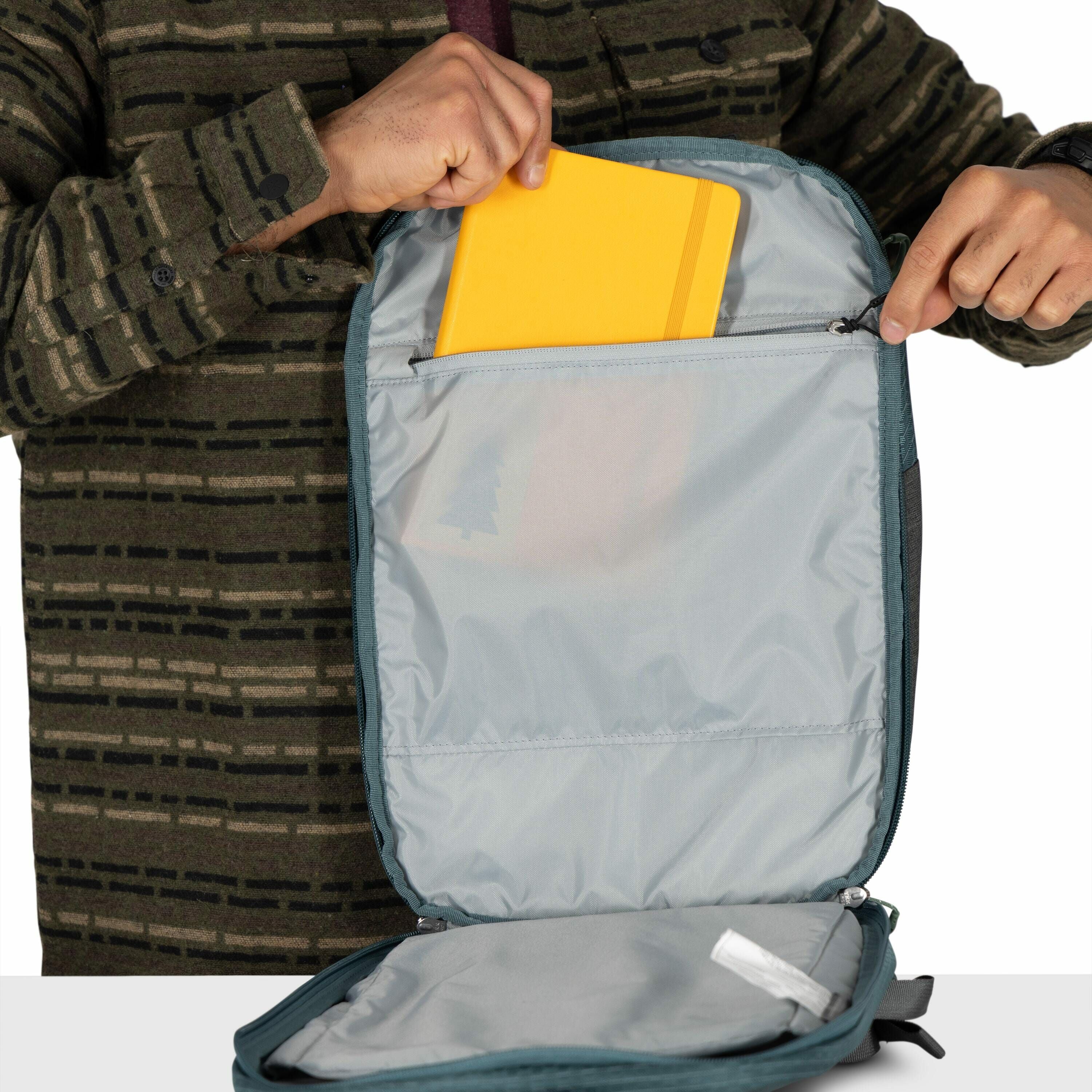 The image shows a person wearing a patterned green jacket, opening a blue-grey backpack. The backpack is open, revealing an inner compartment with a zipper, and the person is placing or removing a bright yellow folder or tablet from this compartment. The