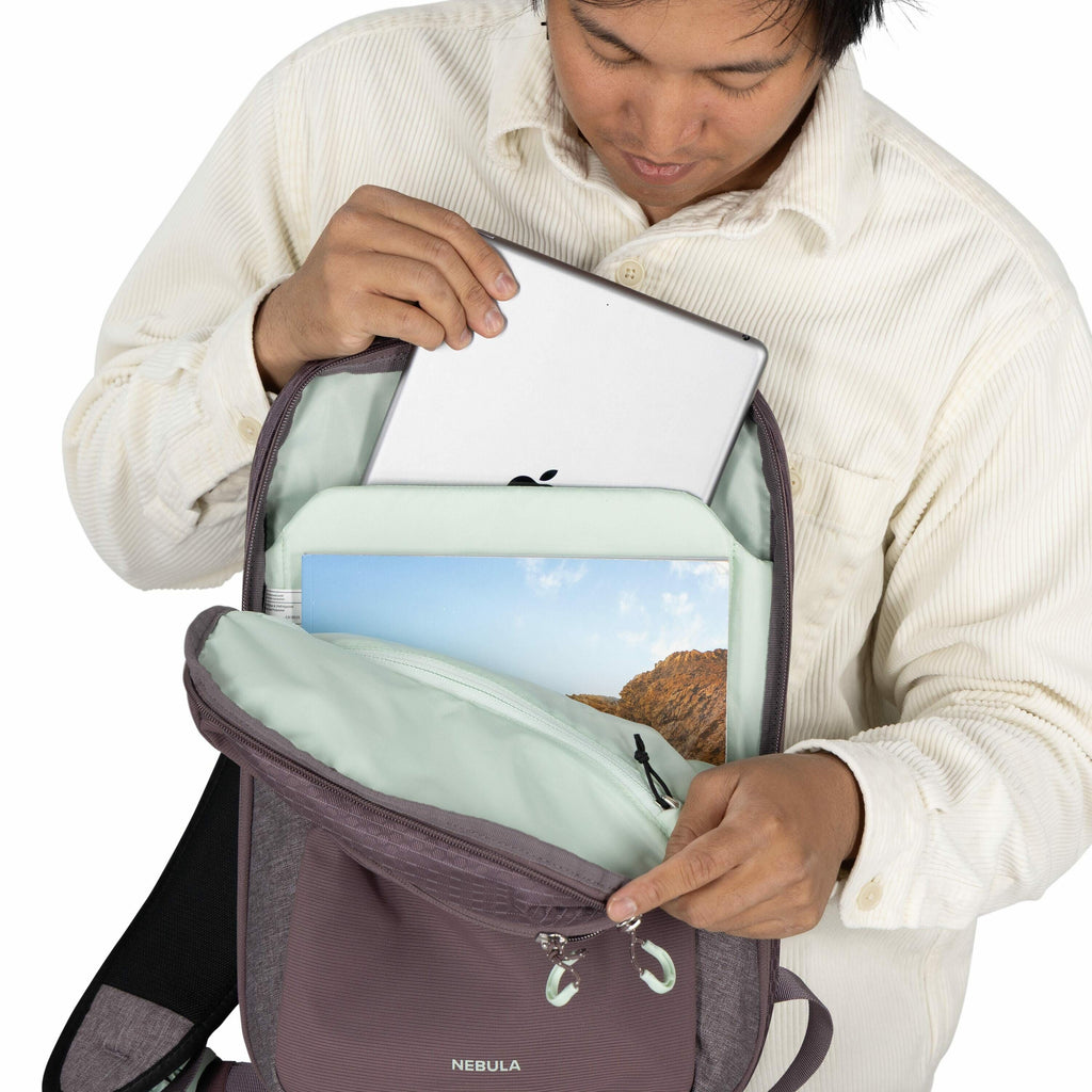 The image shows a person wearing a cream-colored, corduroy button-up shirt holding a backpack. The backpack, branded with the name "NEBULA," is partially open, revealing a neatly arranged interior. Inside the backpack is an Apple device, presumably a lapt