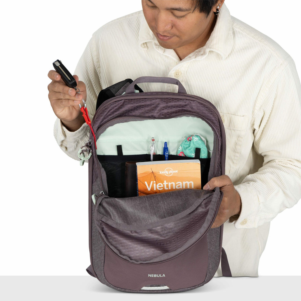 The image features a person holding open a backpack with a grey and purple color scheme. The backpack brand "Nebula" is visible on the front of the bag. Inside the backpack, there are several items organized in pockets: a "Lonely Planet" travel guide labe