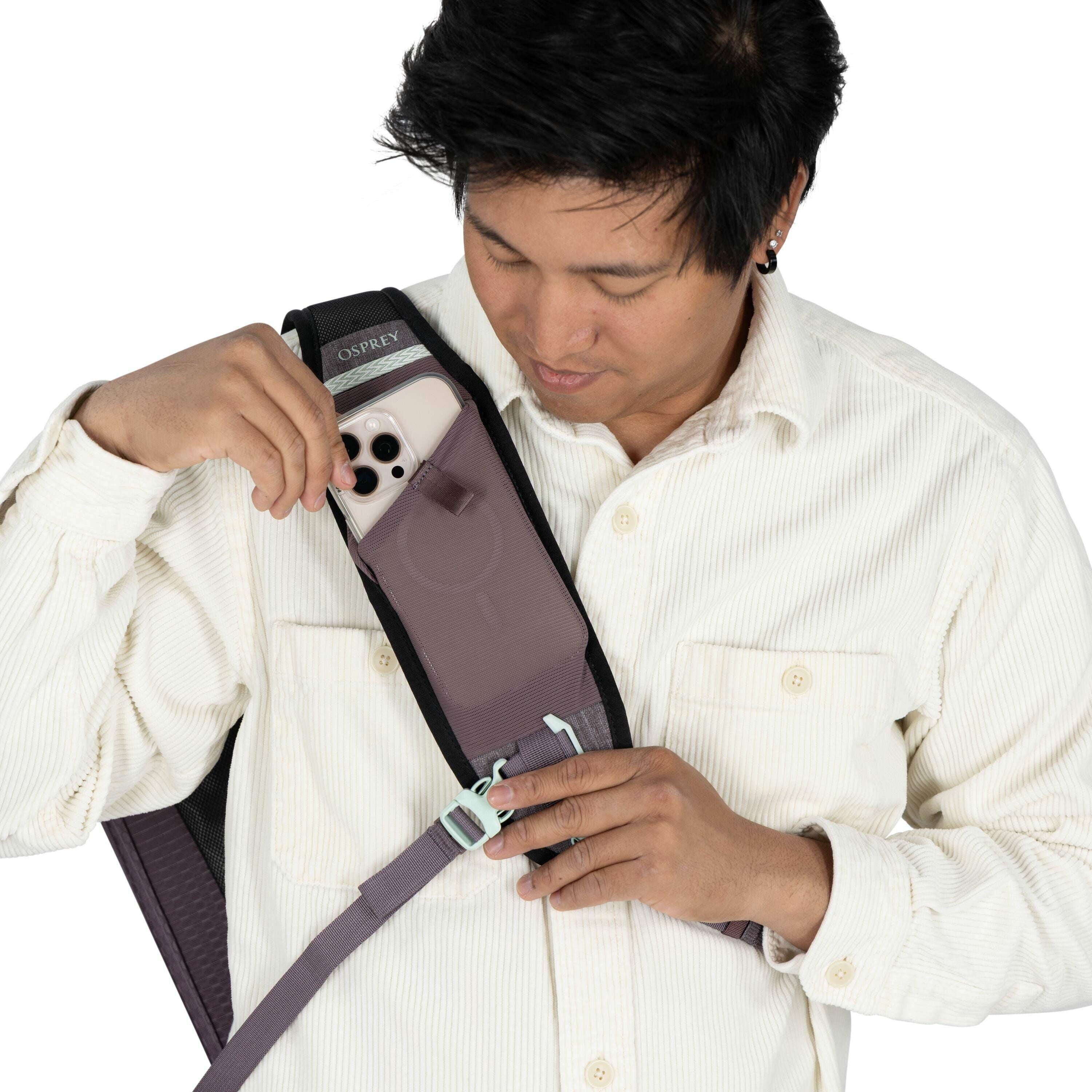 The image shows a person wearing a cream-colored, corduroy button-up shirt, holding a smartphone that is partially inserted into a storage pocket on the shoulder strap of a backpack. The backpack appears to be from the brand Osprey, as indicated by the lo