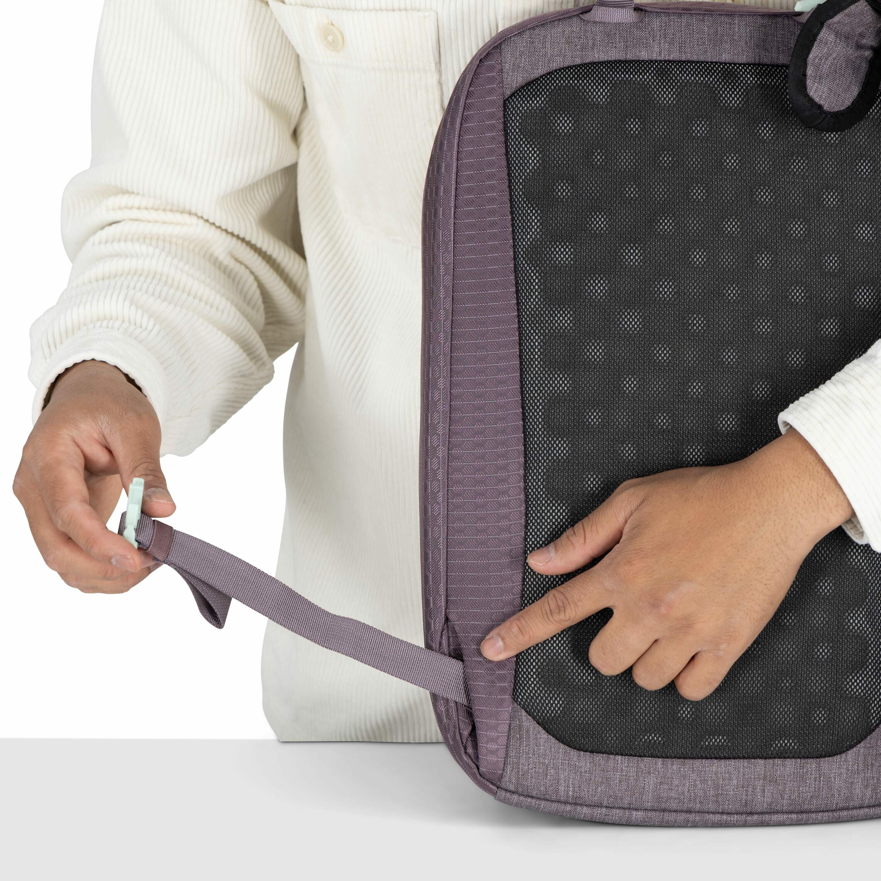 The image shows a person in a cream-colored corduroy shirt adjusting a strap on a backpack. The backpack has a textured black back panel, designed likely for comfort and airflow, with purple fabric outlining the edges. The person's hands are focused on th